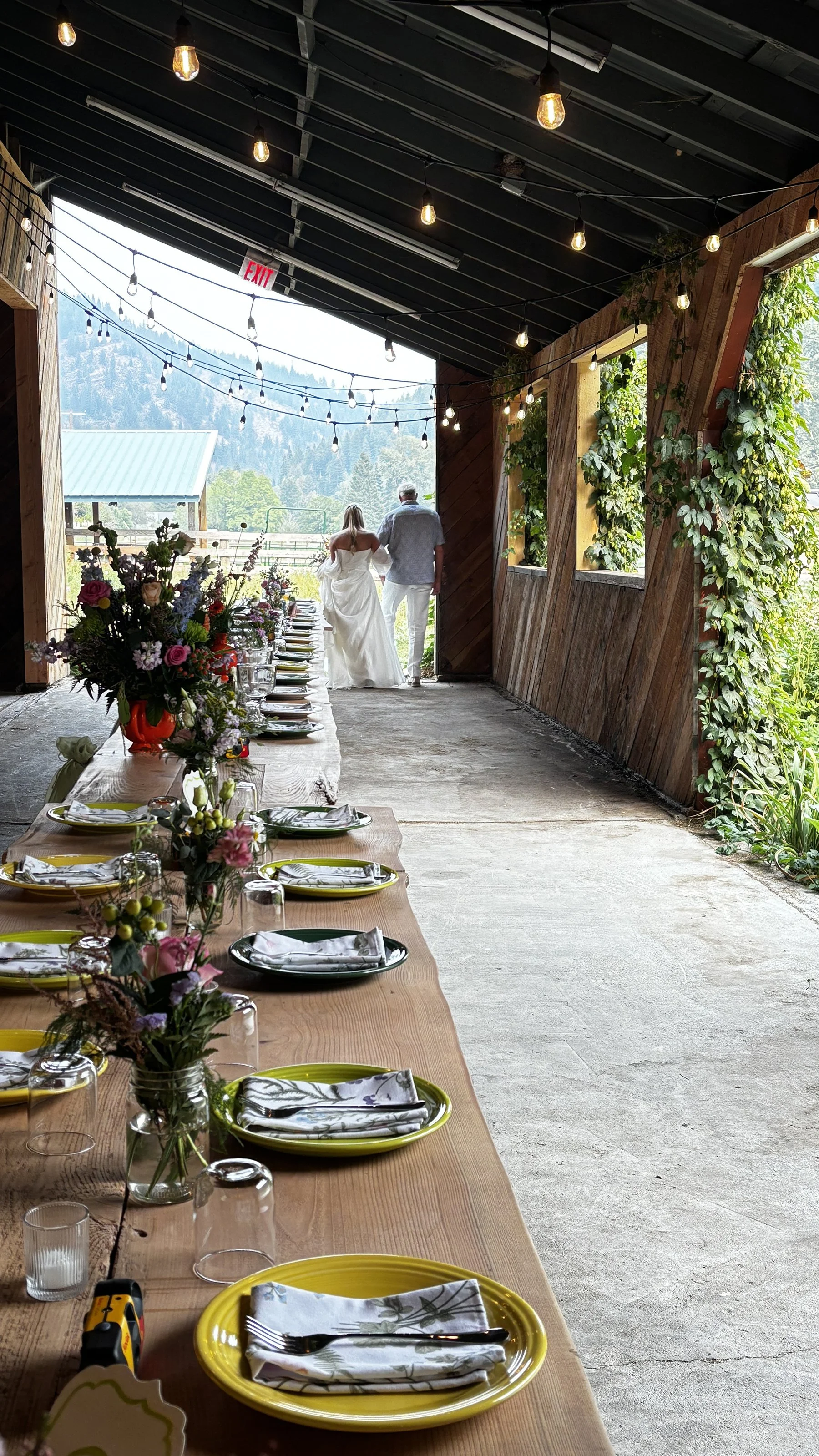 A rustic outdoor dining area with a long wooden table set with yellow plates, floral napkins, glasses, and candles. A large flower arrangement decorates the table. In the background, two people, a bride in a white wedding dress and a man, walk toward a scenic country view with mountains and greenery.