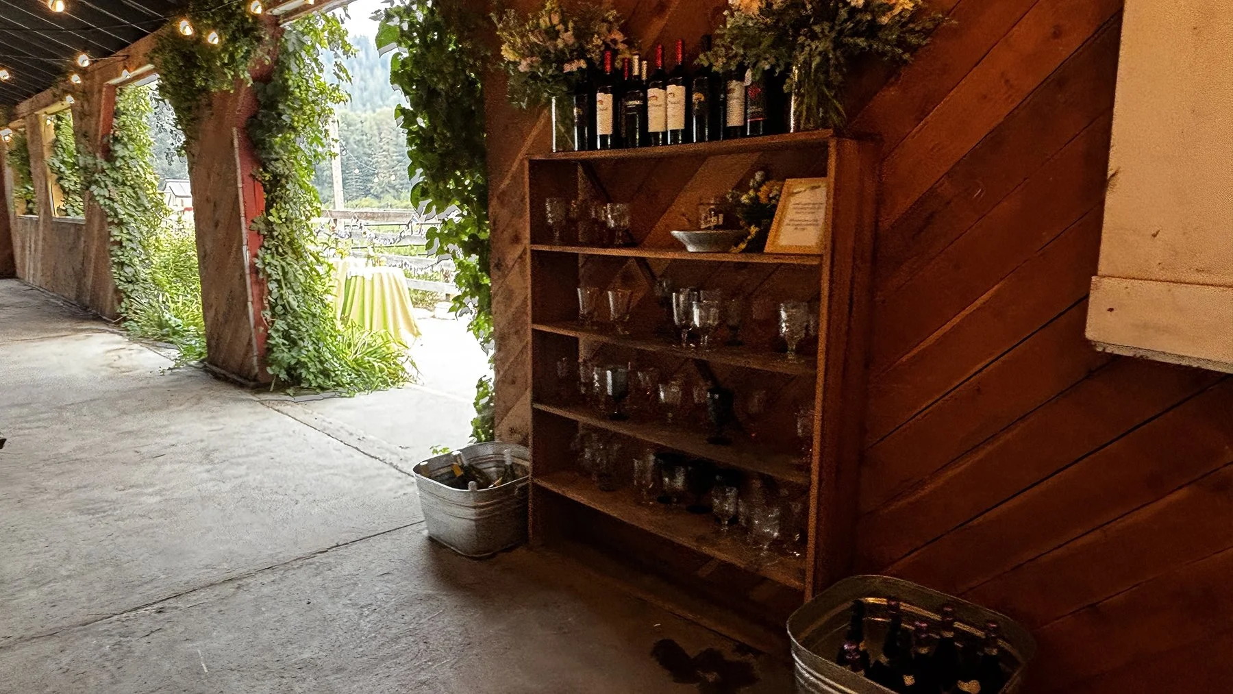 Wooden shelf displaying wine bottles and glassware in a rustic indoor setting, with an outdoor patio visible through open doors.
