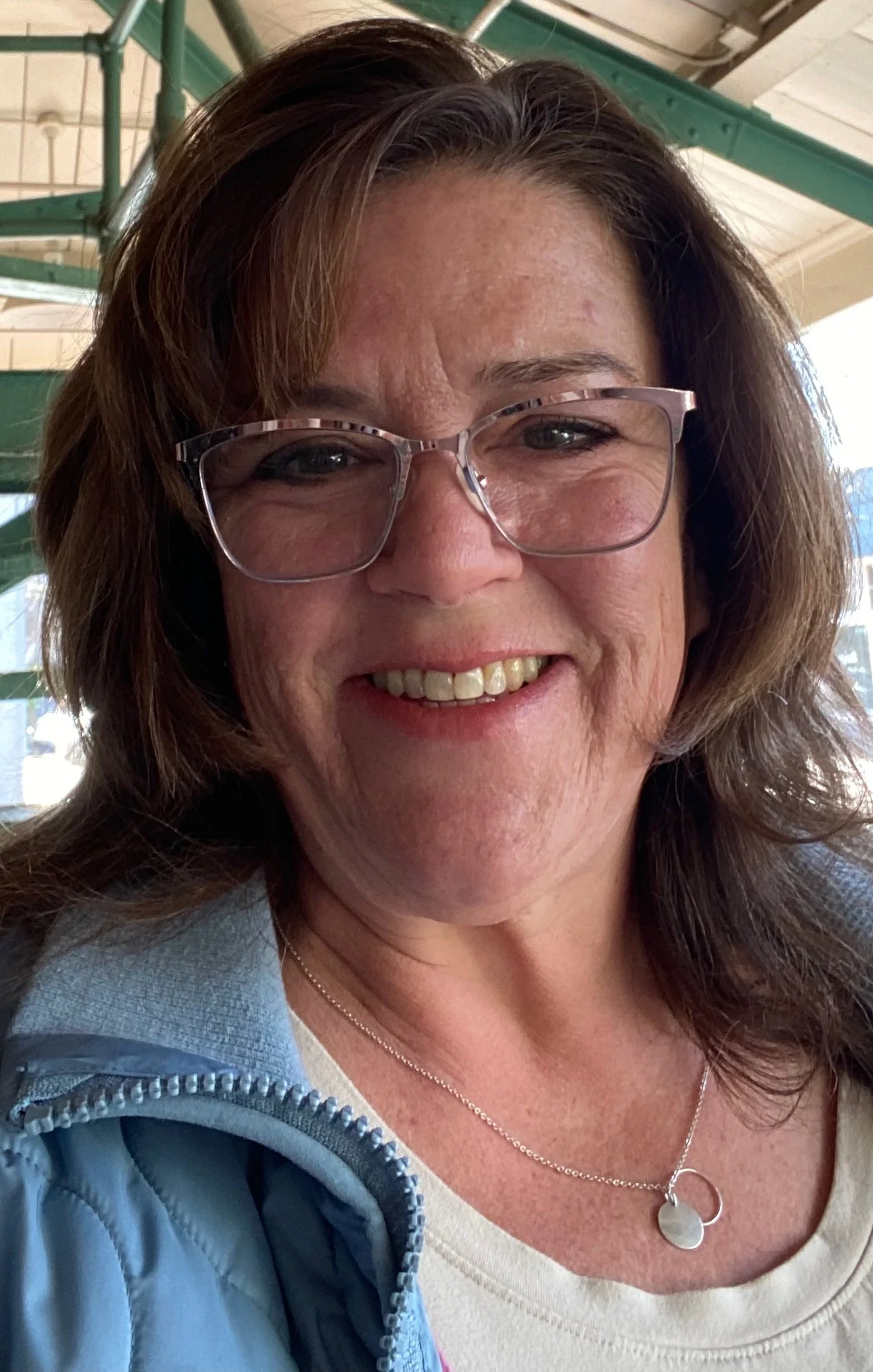 A smiling woman with brown hair, glasses, and a light blue jacket taking a selfie in a sunny outdoor setting.