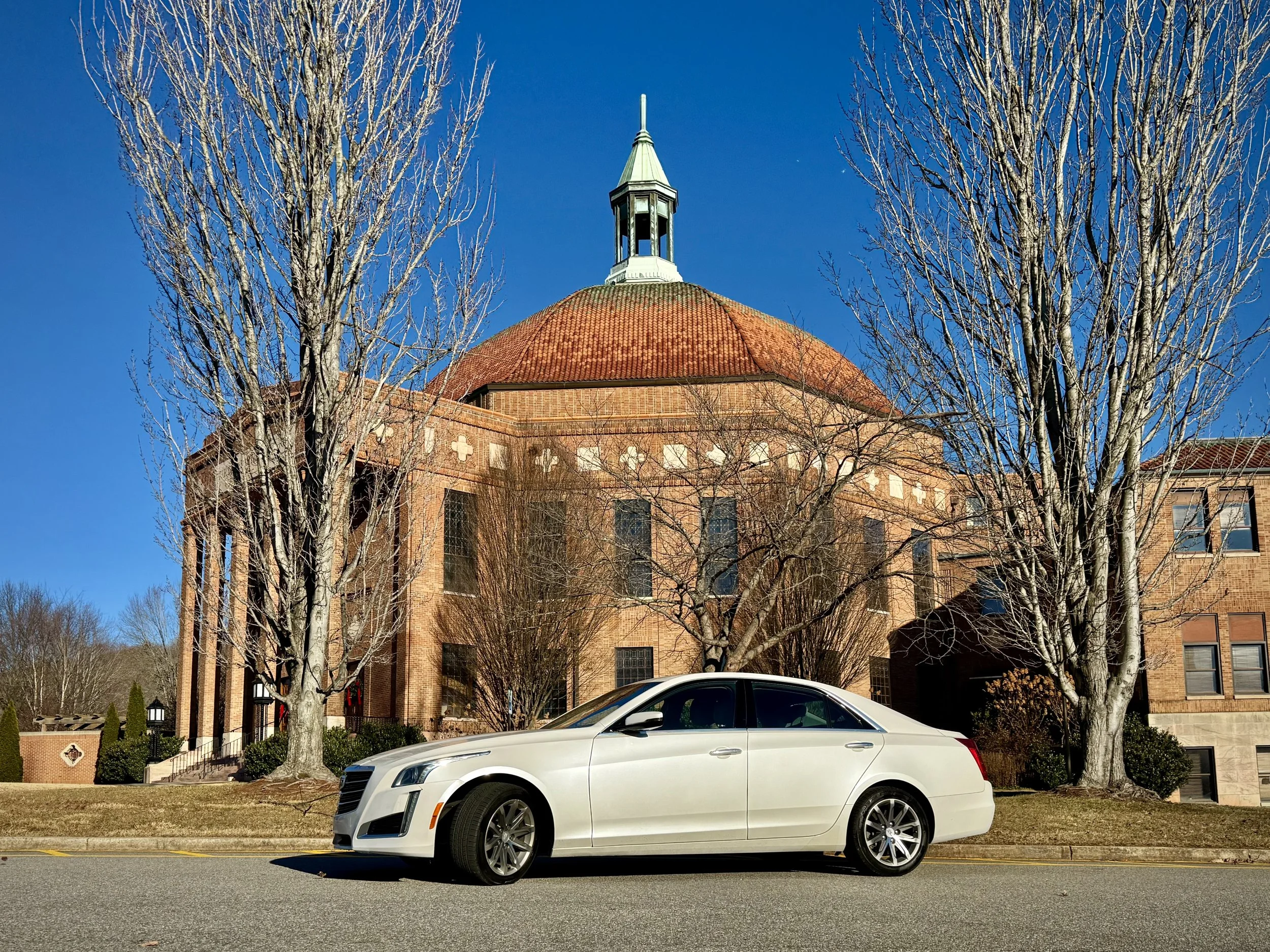 A white luxury car parked on a street in front of a historic brick building with a large dome and a small tower on top, surrounded by leafless trees, under a clear blue sky.