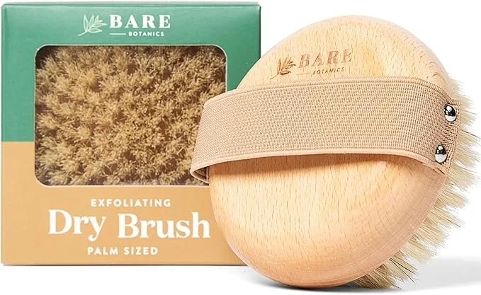 Bare Botanics exfoliating dry brush with a palm-sized wooden handle, beige strap, and bristles, alongside the green and tan packaging box.