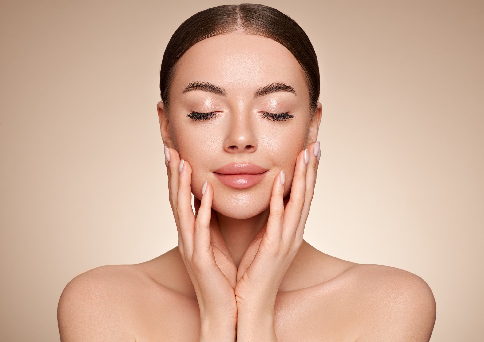 Close-up of a woman with clear skin, gently touching her face with both hands, eyes closed, smiling softly, neutral background.