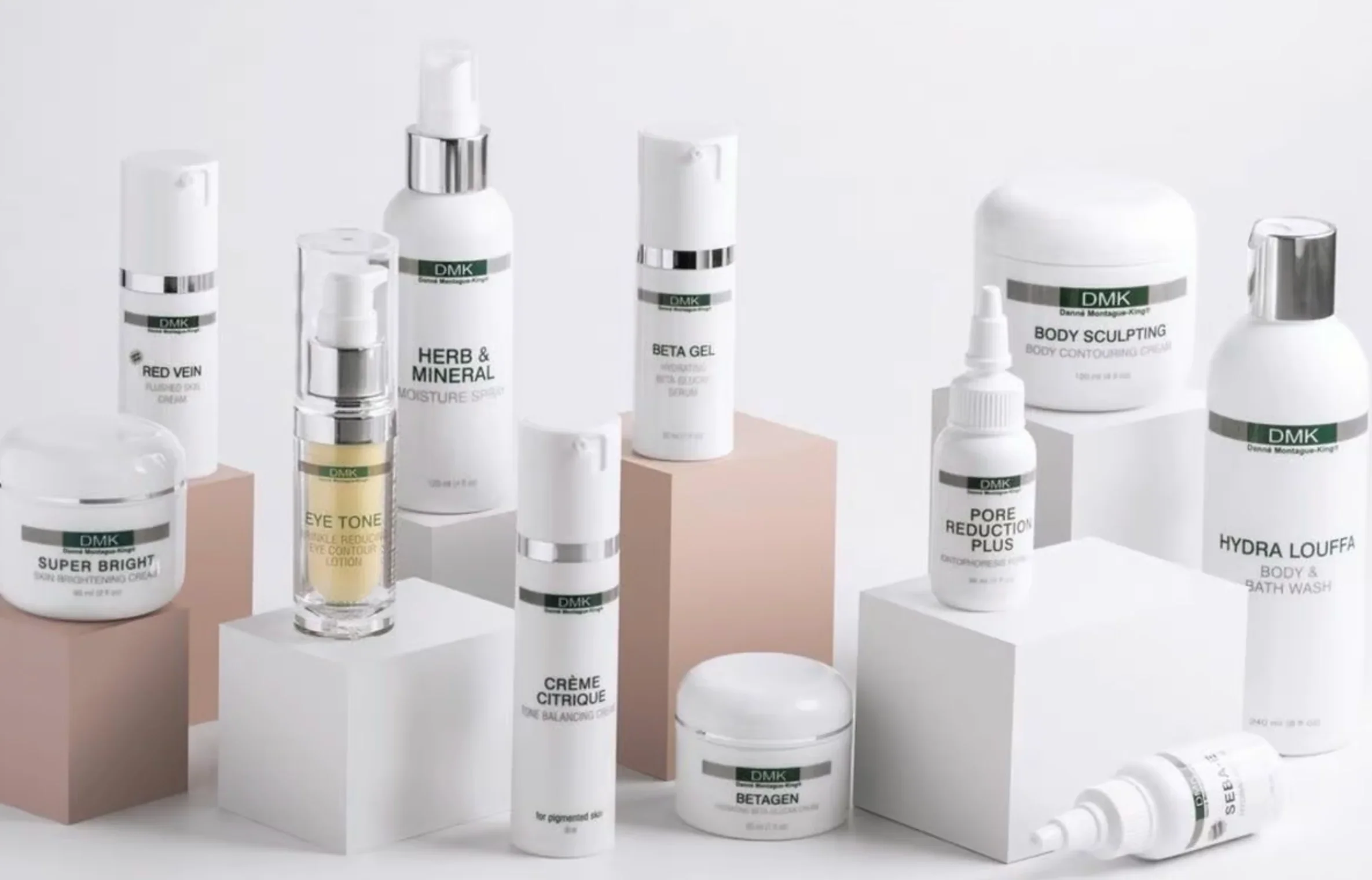 Collection of skincare and body care products on white and pastel-colored blocks, including creams, serums, lotions, and sprays with labels like 'Red Vein,' 'Herb & Mineral,' 'Beta Gel,' 'Super Bright,' and 'Hydra Louffa.'