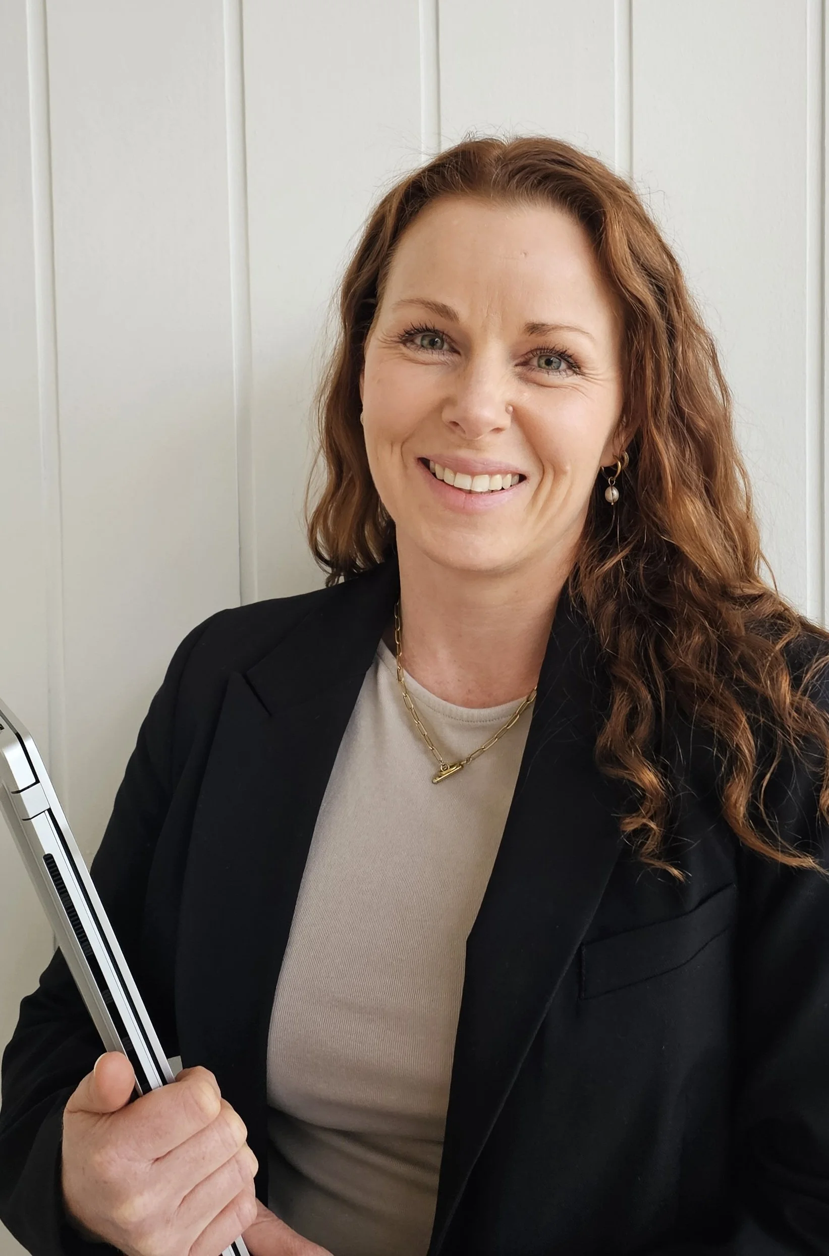 A woman with long, wavy red hair smiling, wearing a black blazer and beige top, holding a closed laptop, standing against a white paneled wall.