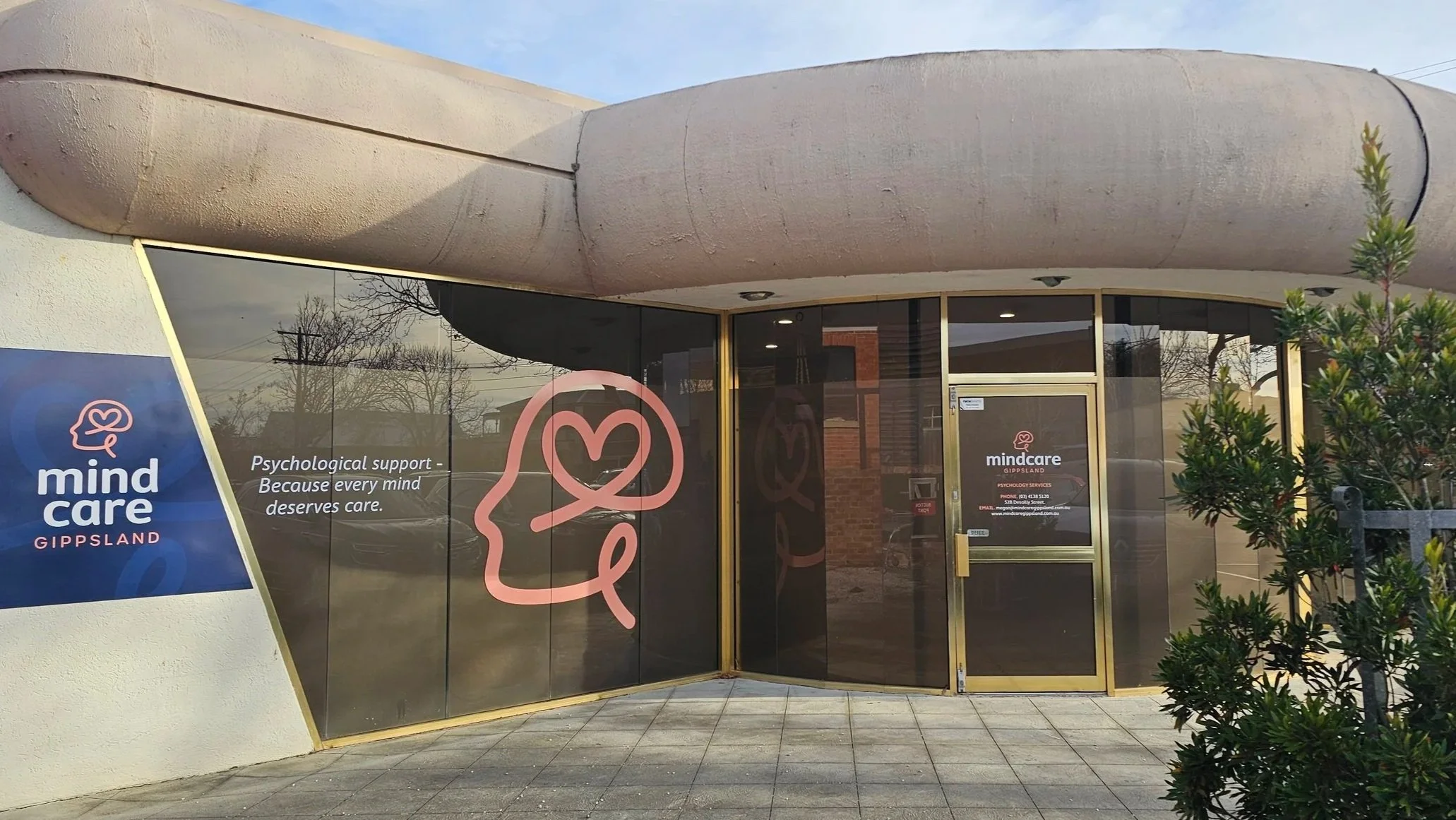 Facade of a psychology practice named 'MindCare Gippsland,' with a peach coloured line art logo of a head with a heart as the brain, and glass doors with signs.