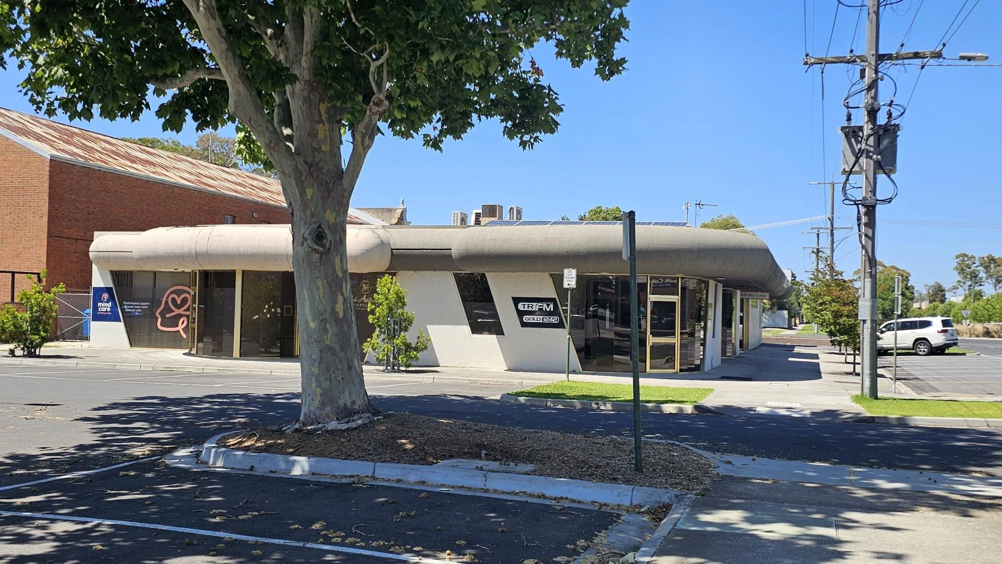 A single-story building with a curved roof, large glass windows and doors, and street parking in front, surrounded by trees and utility poles on a sunny day.