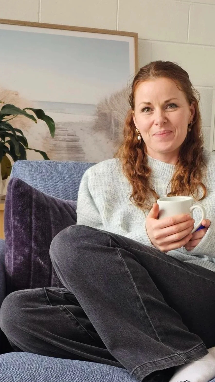 A woman with curly red hair sitting on a couch, holding a white mug, wearing a light gray sweater and dark jeans, in a cozy room with a large plant and a framed picture in the background.