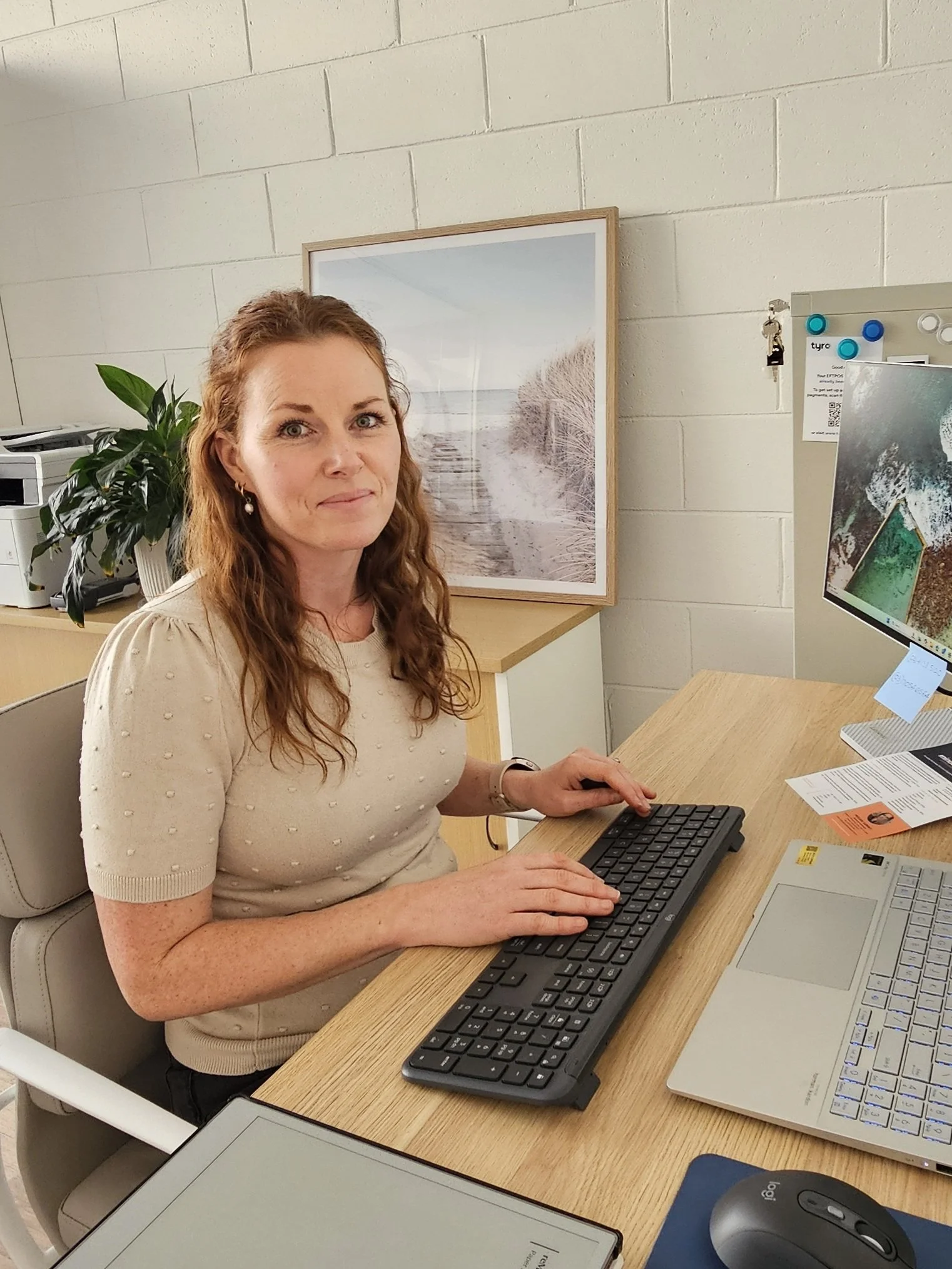 A woman sitting at a desk with a keyboard, mouse, and a computer monitor, in an office with framed beach photo on the wall and a plant.