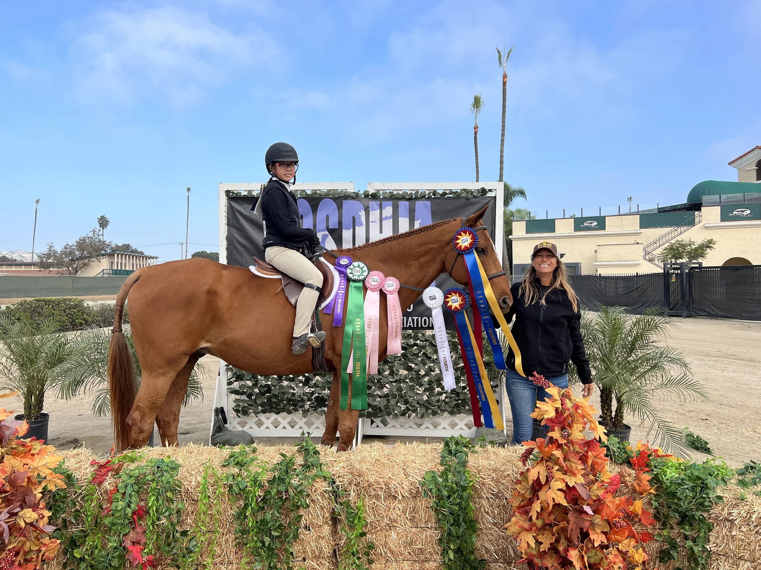 A young girl riding a brown horse decorated with colorful ribbons, standing on a hay bale, with a smiling woman standing beside the horse holding flowers. They are outdoors with palm trees and buildings in the background, celebrating an equestrian event.