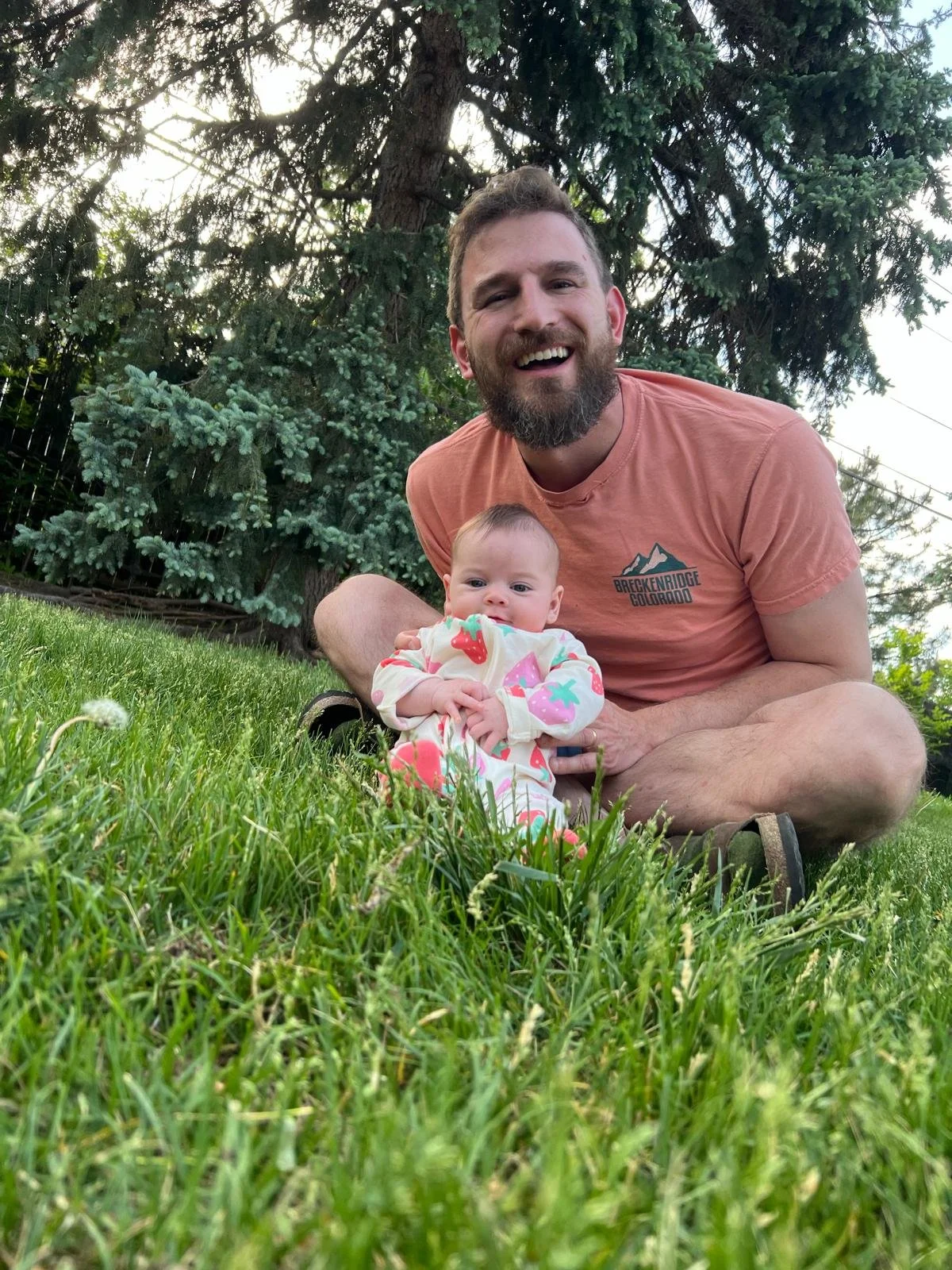 A man with a beard and a smiling face, wearing a salmon-colored T-shirt, is sitting in the grass outdoors, holding a baby girl who is lying on her stomach on the grass. The baby is wearing a colorful onesie with pink, green, and orange patterns. The background includes large trees and a wooden fence.