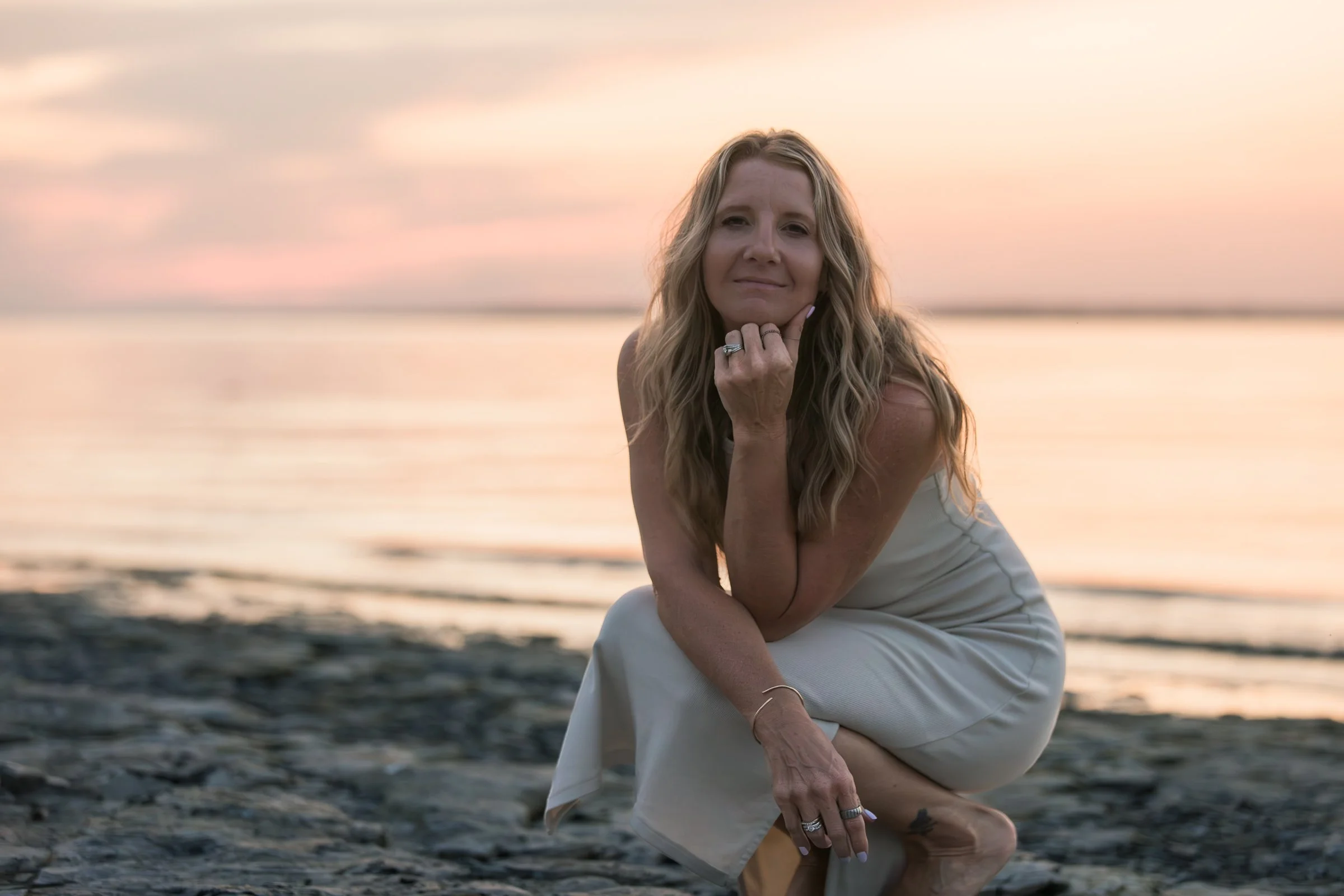 A woman with long blonde hair sitting on a sandy beach during sunset, wearing a white dress, with a serene expression on her face.
