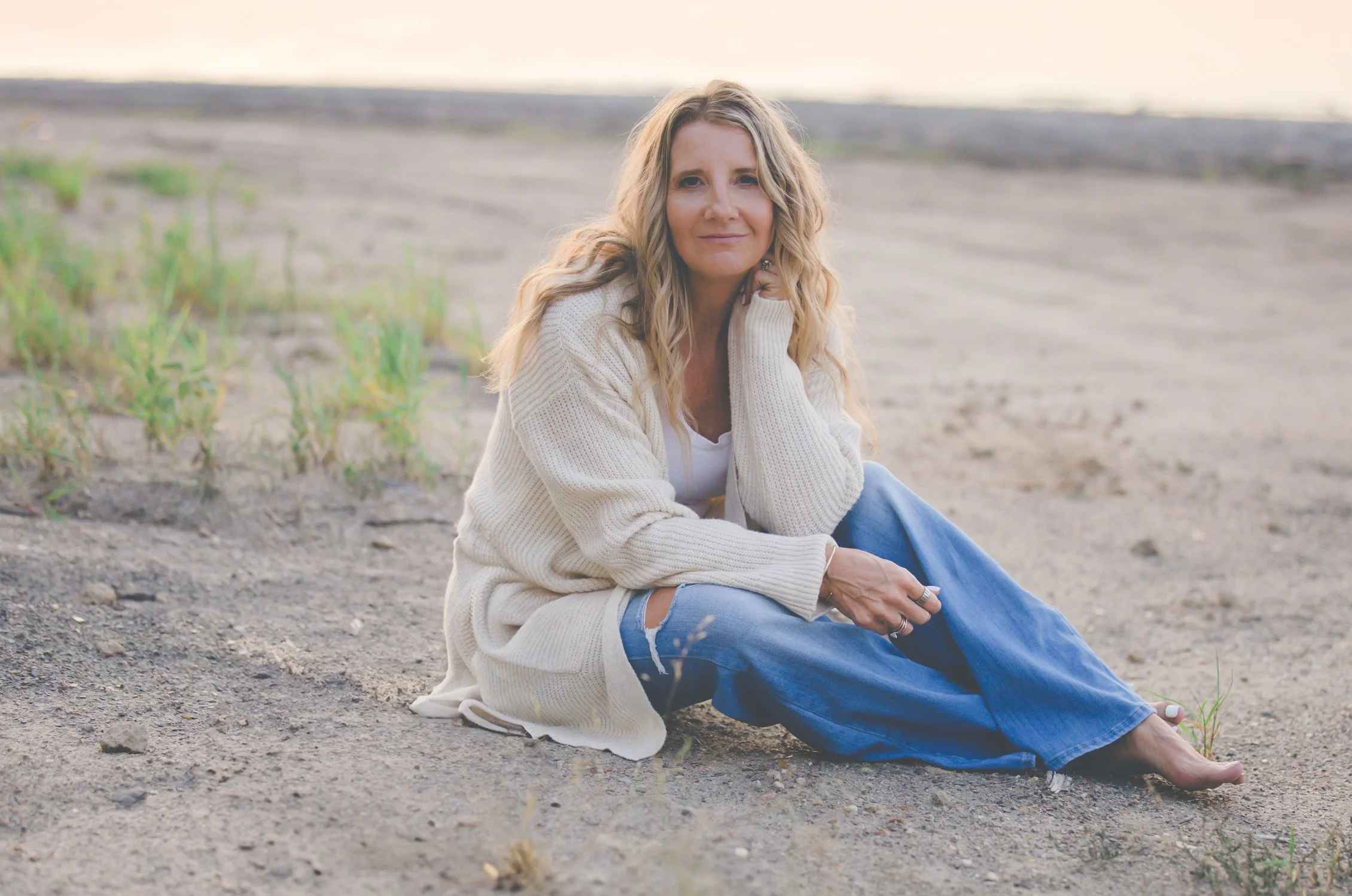 A woman with long blonde wavy hair, wearing a white sweater and ripped blue jeans, sits on a sandy beach with sparse green grass, looking at the camera with a gentle smile, during sunset or sunrise.