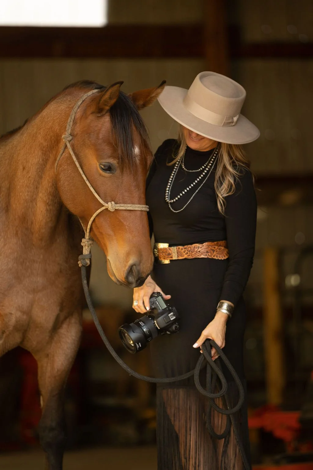 A woman in a large beige hat, black dress, and pearl necklaces holding a camera and standing beside a brown horse with a beige halter.