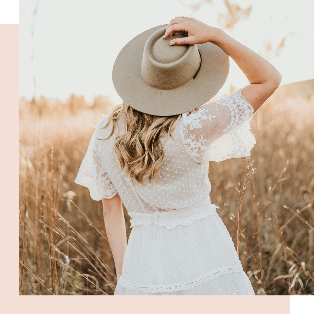 Woman in white lace dress holding wide-brimmed hat, standing in a golden field at sunset