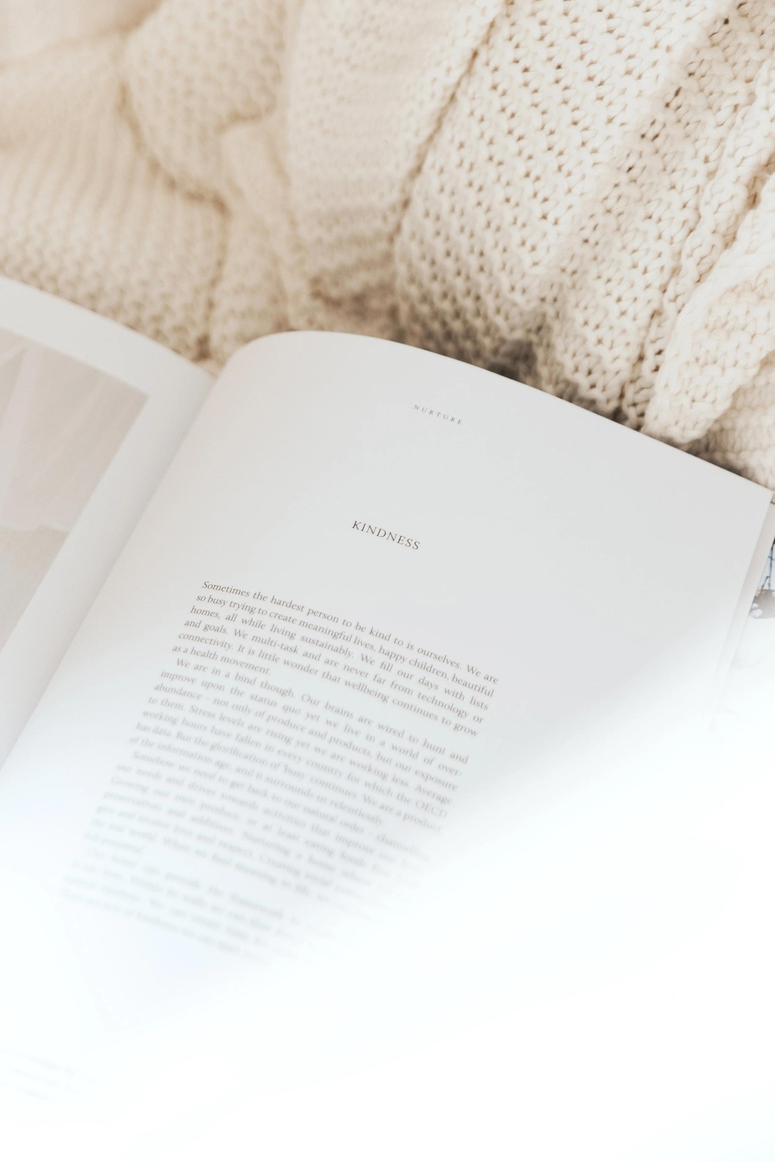 An open book displaying a page with the title 'NURTURE' and the subtitle 'KINDNESS' on a surface with soft knitted cream-colored fabric in the background.