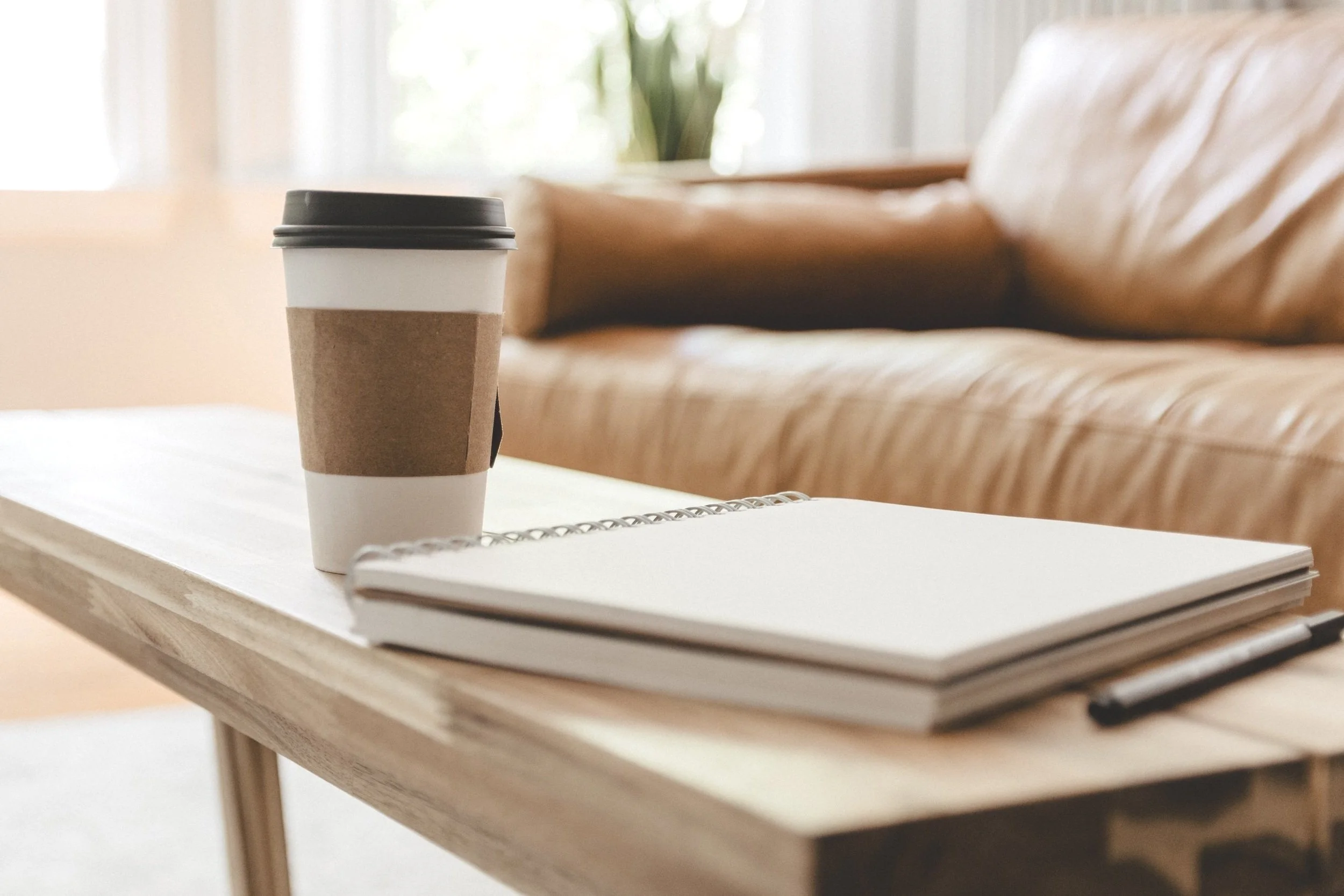 A disposable coffee cup with a black lid on a light wooden table, a spiral notebook and pen nearby, in a cozy room with a couch and a large window with curtains in the background.