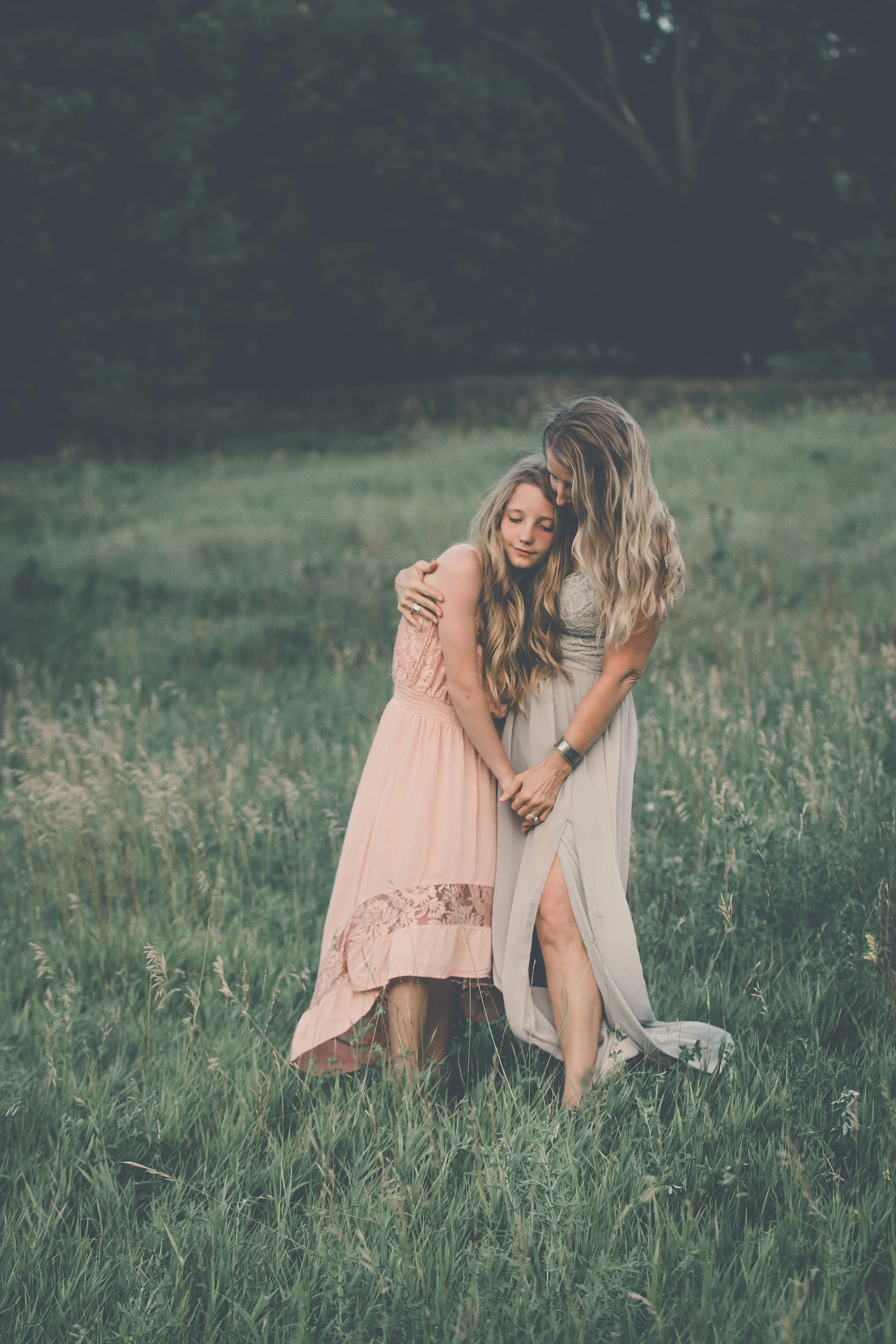 Two women hugging each other in a grassy field during sunset with trees in the background.