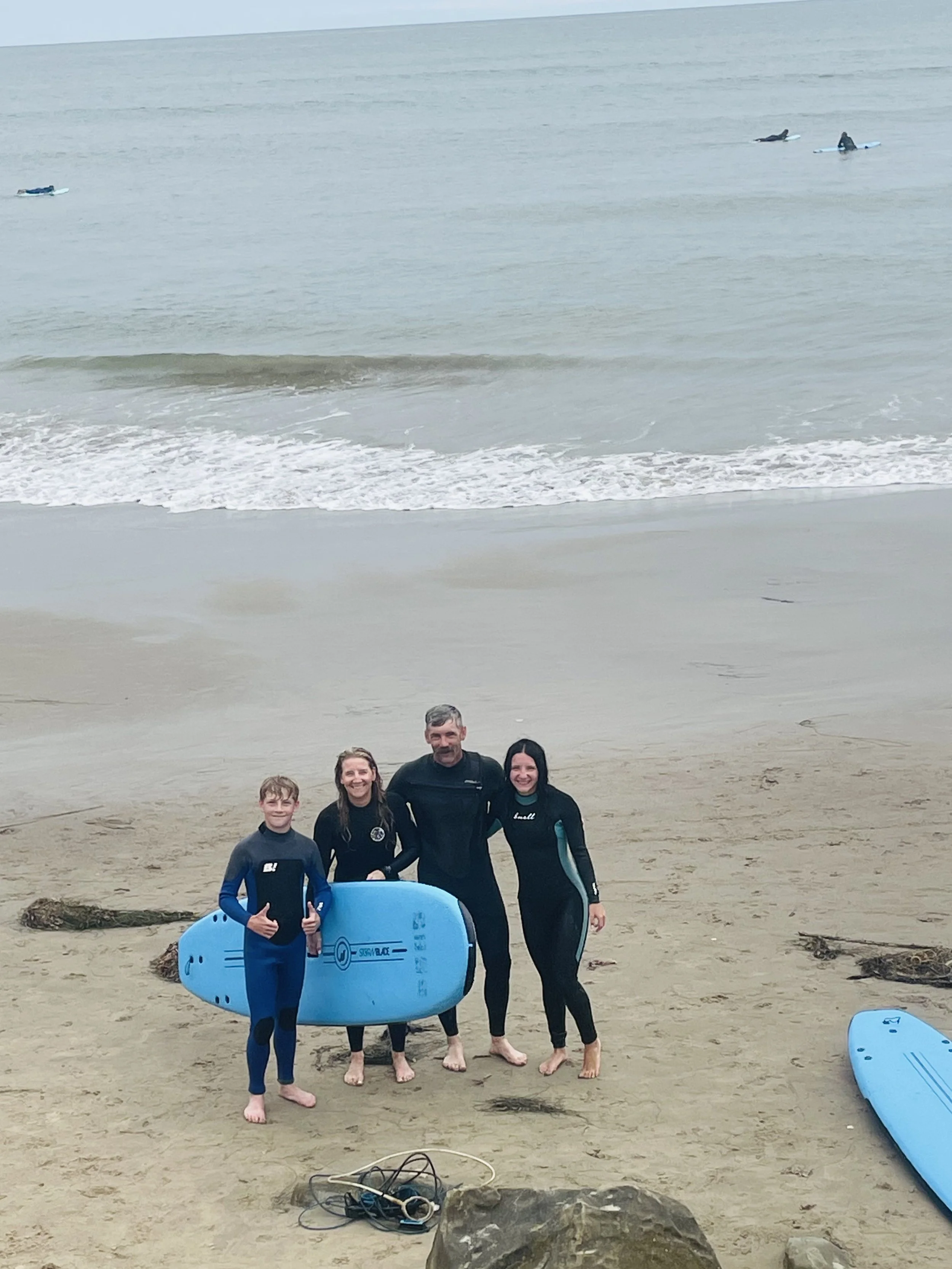 Group of five people, including children and adults, standing on the beach with surfboards, smiling for the camera, with ocean waves and cloudy sky in the background.