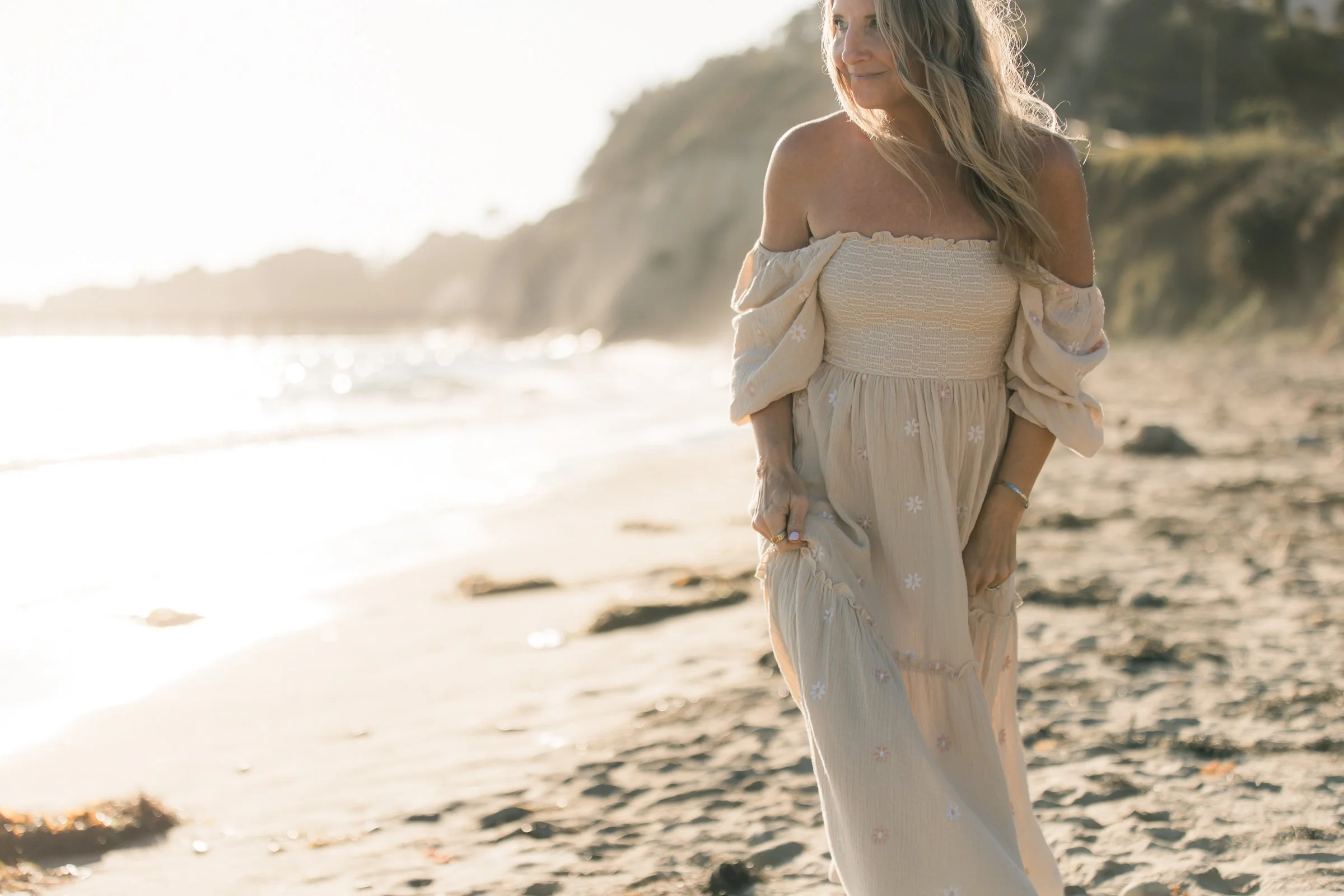 A woman with long, blonde hair wearing a flowing beige dress with white floral patterns, standing on a sandy beach during sunset, with cliffs and ocean in the background.