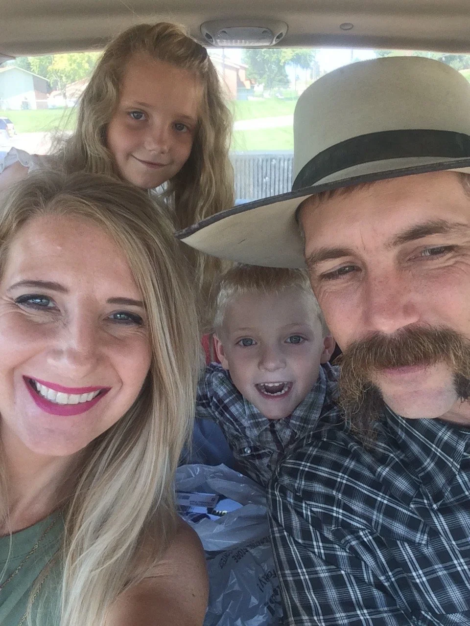 Family of five taking a selfie inside a vehicle. The woman has blonde hair and is smiling, the man has a large mustache and is wearing a cowboy hat, the girl has long wavy blonde hair, and the two young boys have short blonde hair. All are smiling or making expressions, and the background shows a suburban neighborhood through the vehicle window.