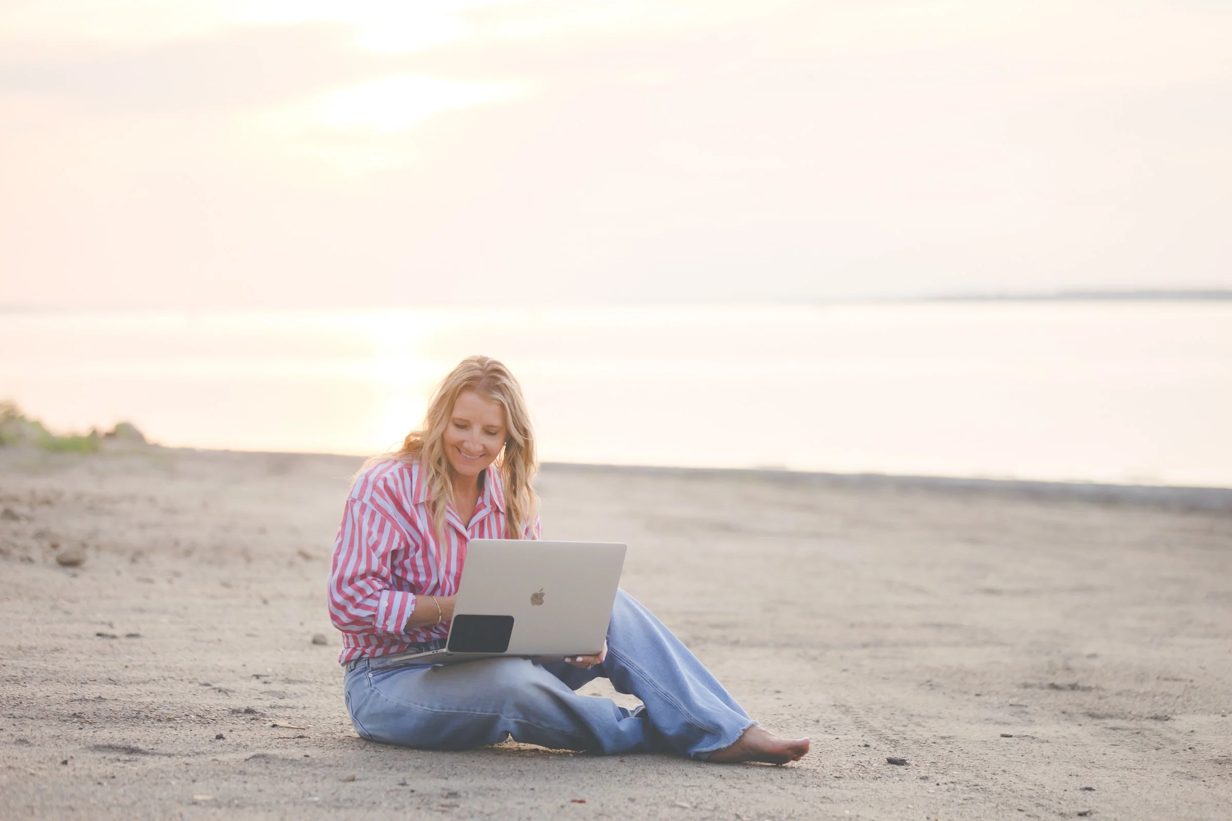 A smiling woman sits on the sandy beach using a laptop, wearing a red and white striped shirt and blue jeans, with the ocean and sky in the background.