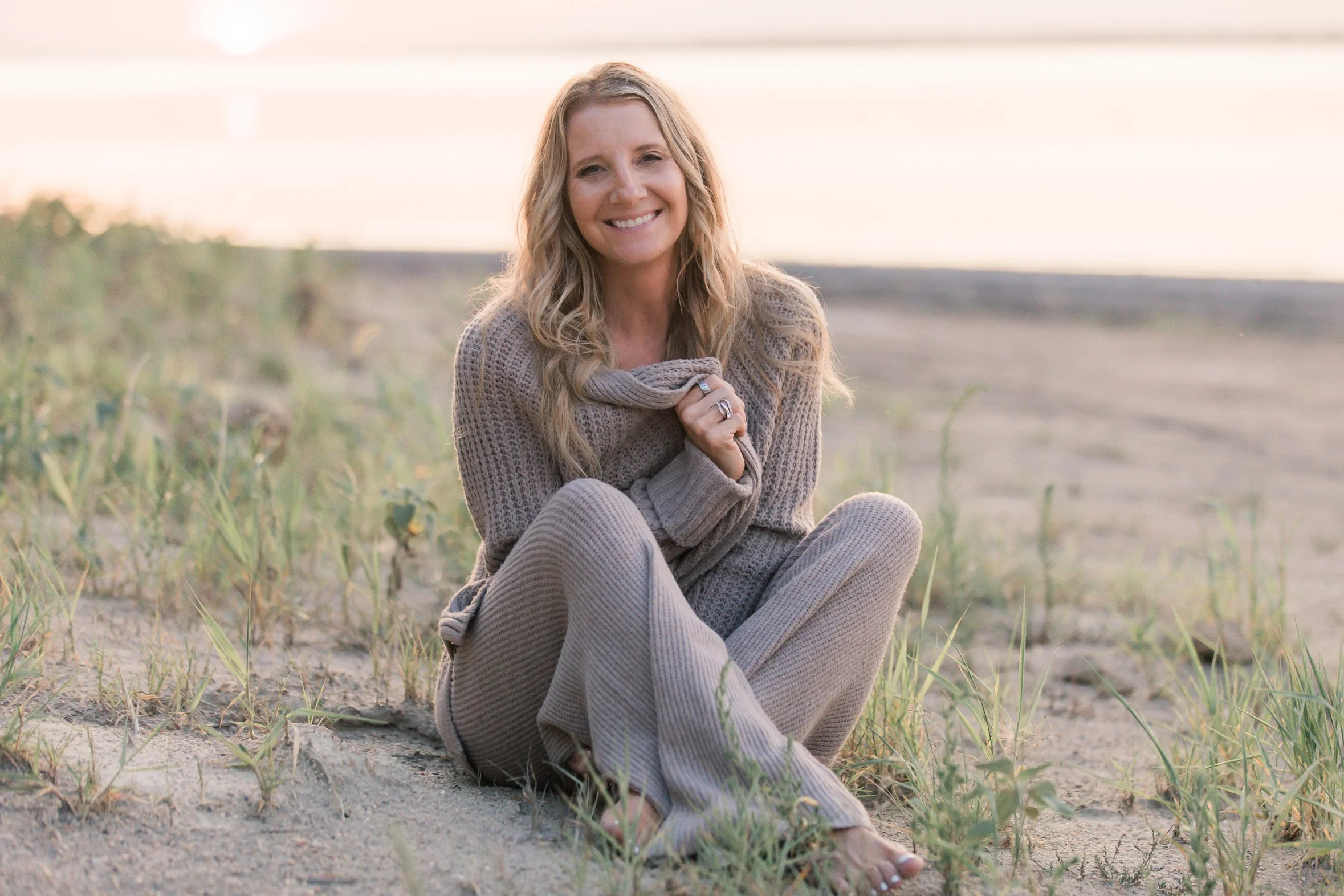 A woman with long blonde hair smiling while sitting on the sand at the beach, holding her sweater close to her chest, with sea grasses and a blurry ocean in the background during sunset.
