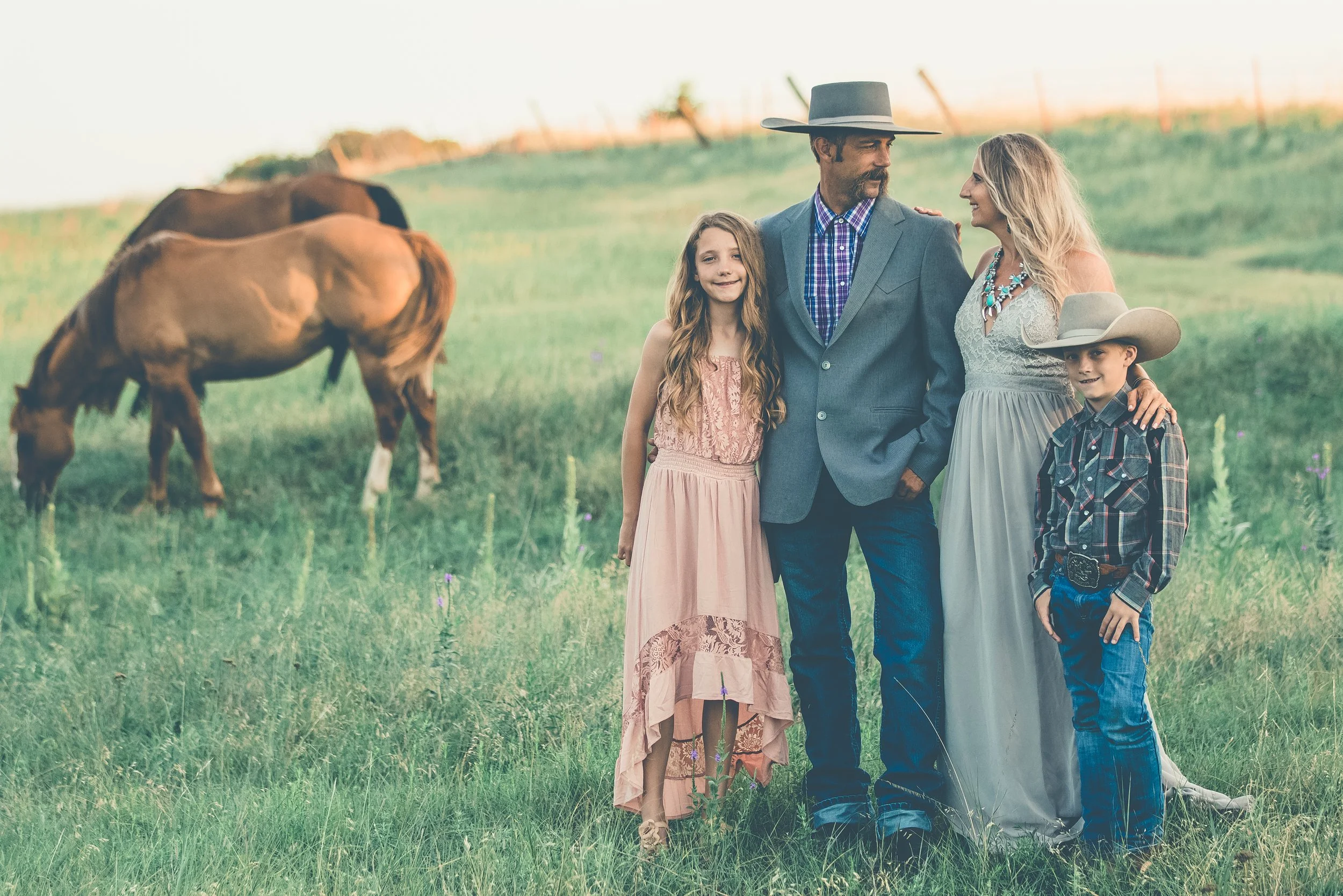 A family of four standing together in a grassy field with horses grazing nearby, with a fence and a sunset sky in the background.