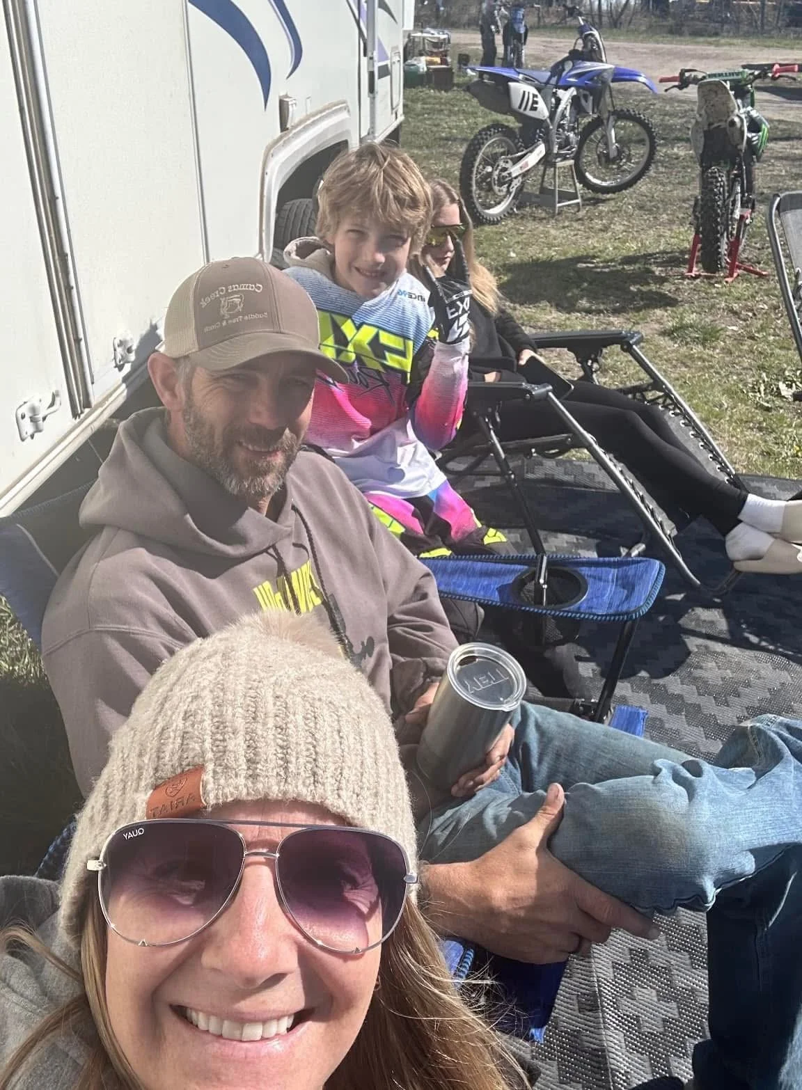 Group of people sitting outside near motorcycles, dirt bikes, and an RV, enjoying a sunny day.