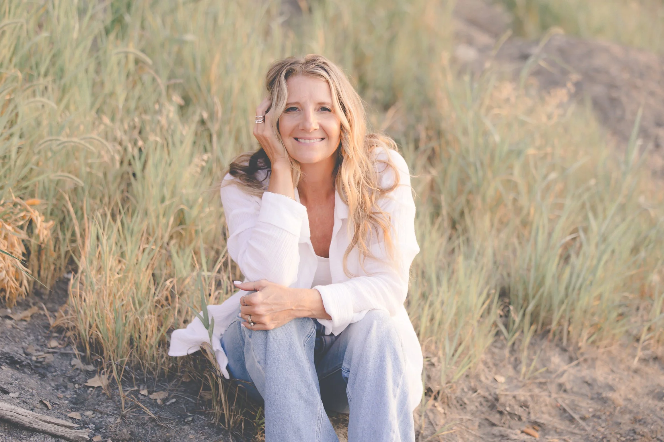 A woman with blonde hair sitting outdoors near tall grass, smiling at the camera, wearing a white shirt and light blue jeans.