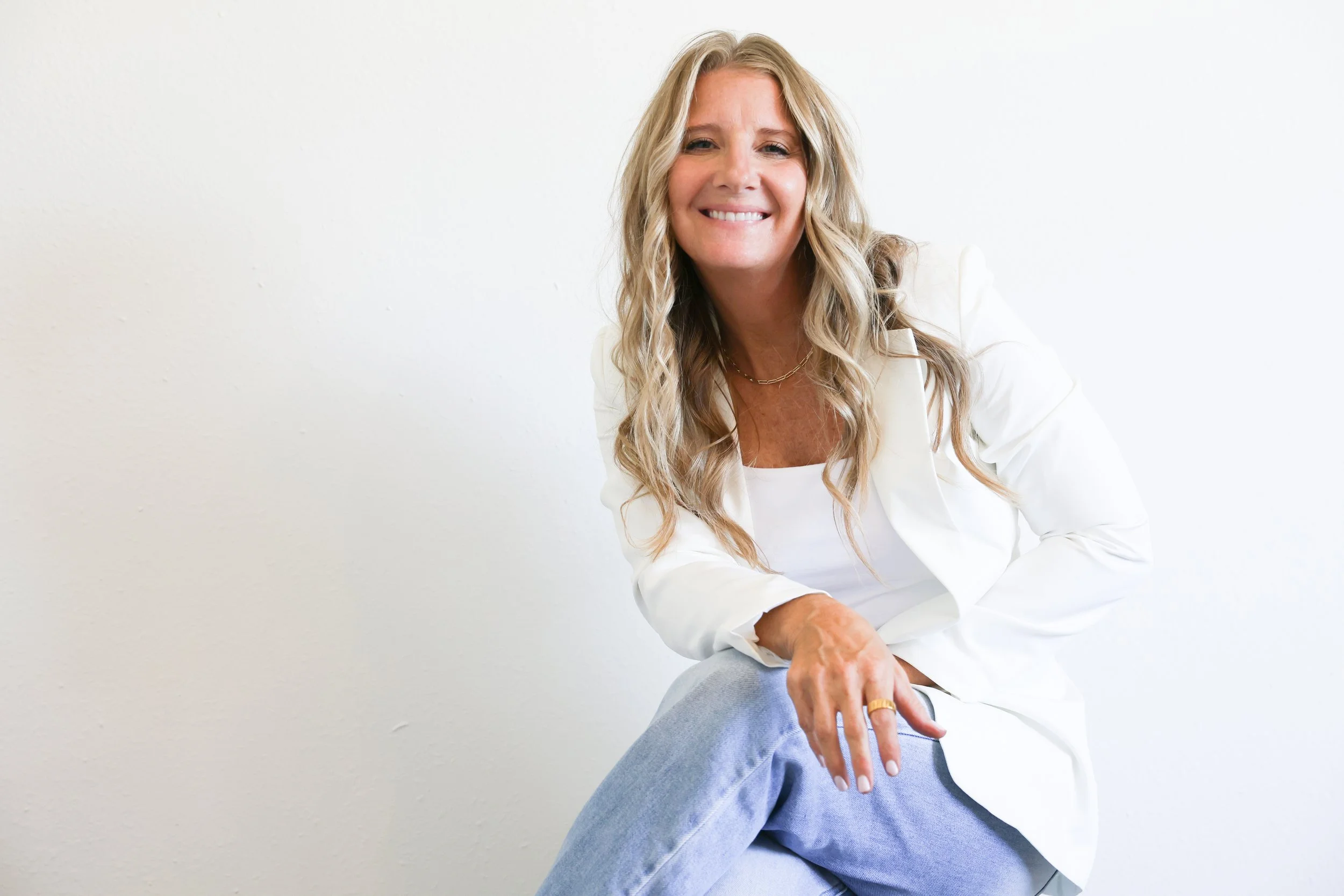 A woman with long blond hair smiling, sitting against a white wall, wearing a white blazer, a gold necklace, and light-colored jeans.