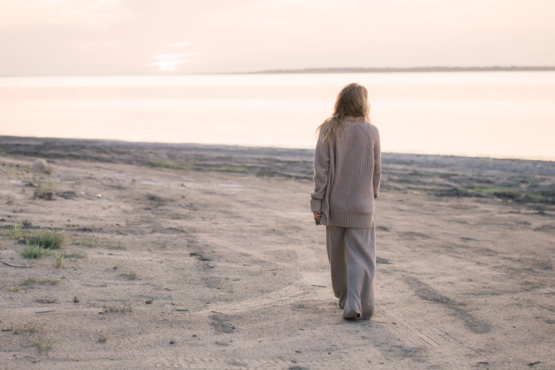 A person with long hair wearing a beige coat and beige pants walks on a sandy beach near water during sunset.