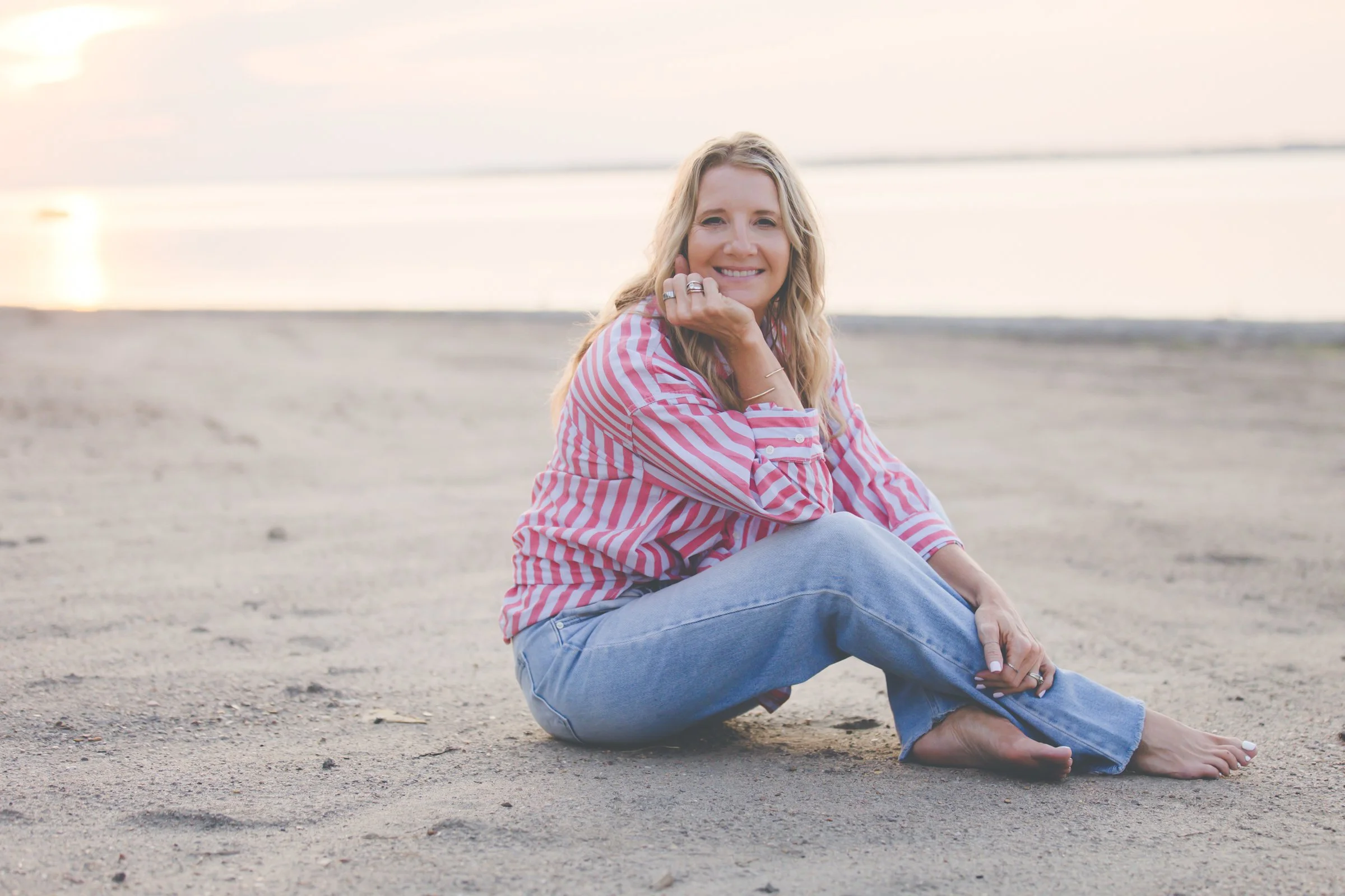 A woman with blonde hair, wearing a pink and white striped shirt and blue jeans, sitting on the sandy beach with her legs crossed and smiling at the camera during sunset.