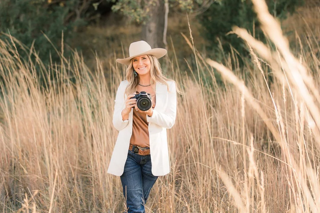 A smiling woman in a white hat and blazer holding a camera in a field of tall grass.