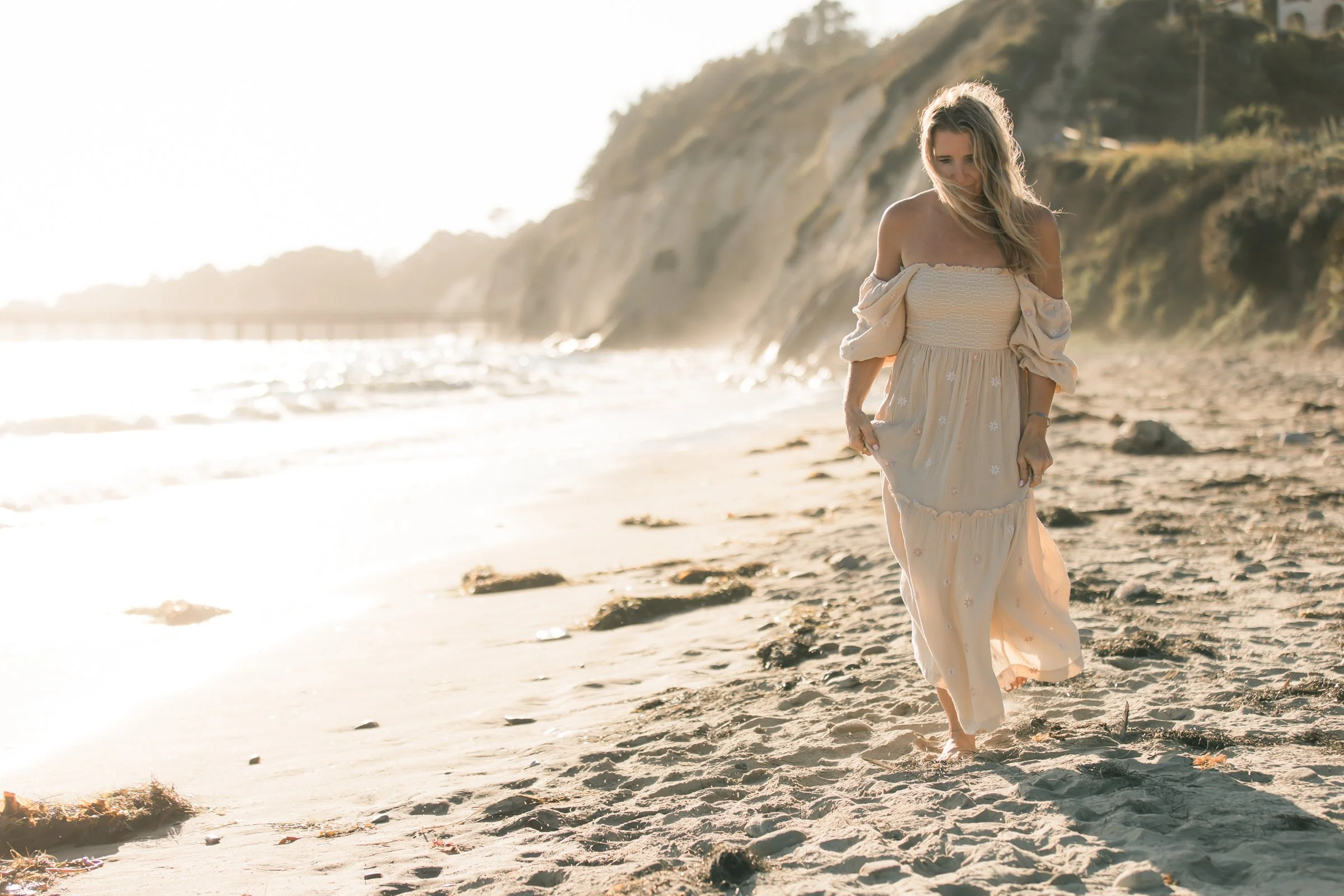 A woman in a flowing dress walks along a beach with cliffs in the background, illuminated by the setting sun.