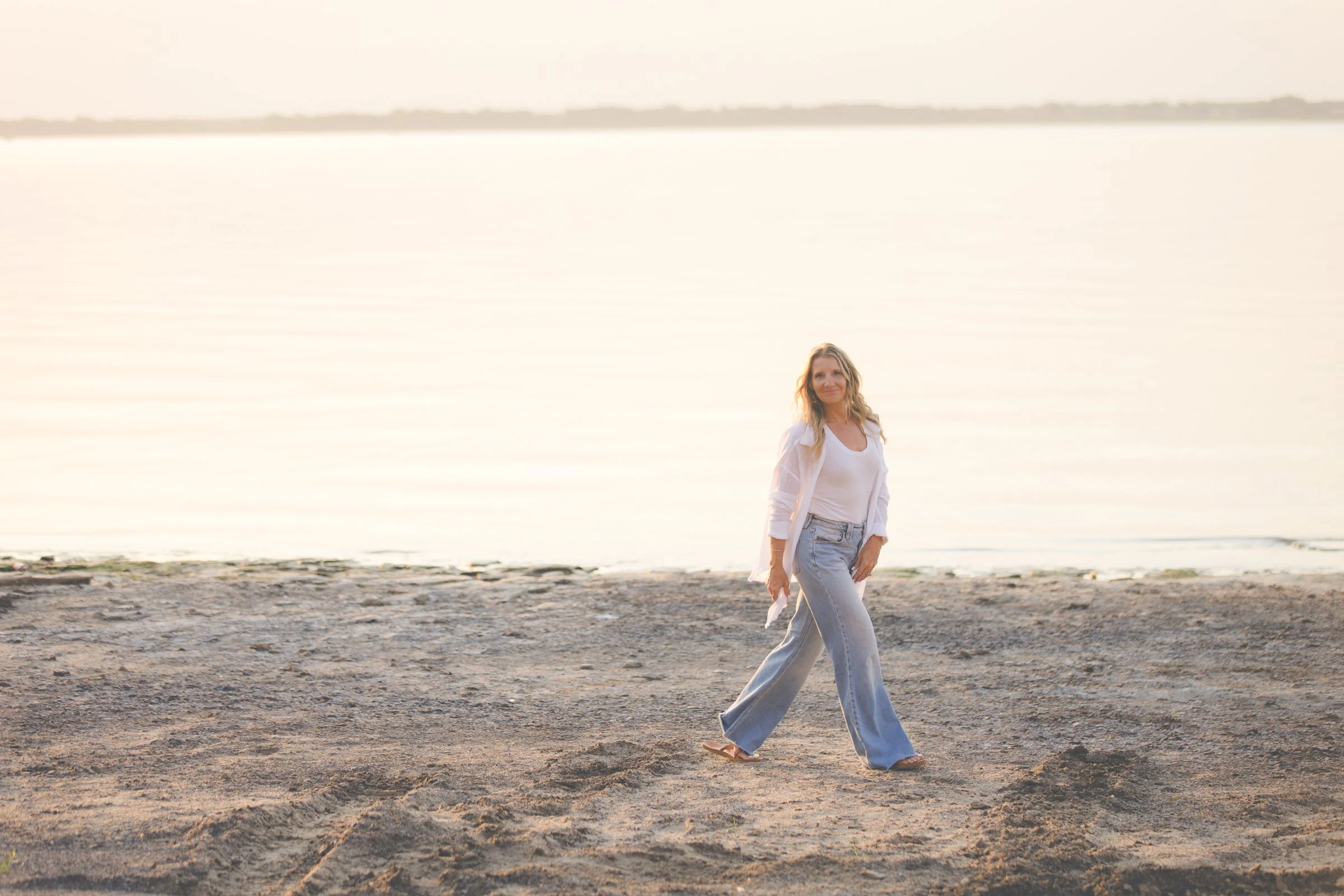 A woman walking on a sandy beach with the ocean and sky in the background, smiling and dressed casually in a white top, light jacket, and wide-leg jeans.