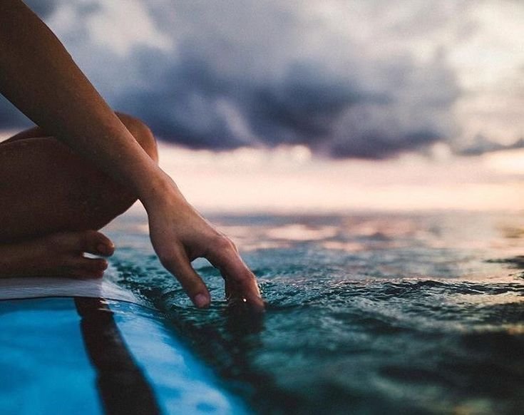 A person's hand touching the water on a surfboard at the ocean during sunset or sunrise.