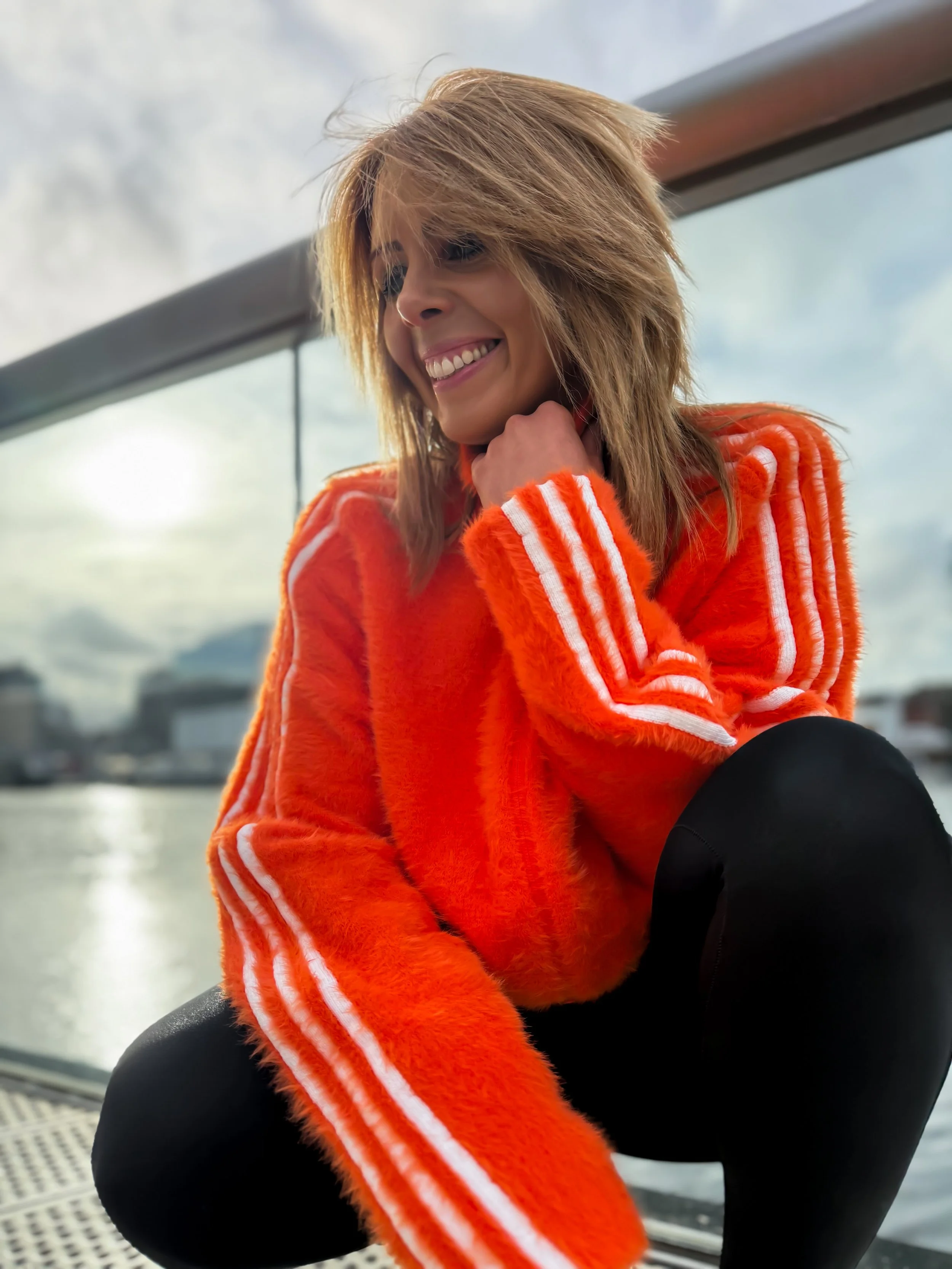 A woman with blonde hair in a trendy haircut, smiling and looking to the side, dressed in a bright orange, fuzzy athletic jacket with white stripes down the sleeves, sitting near a large window with a scenic view of water and cloudy sky.