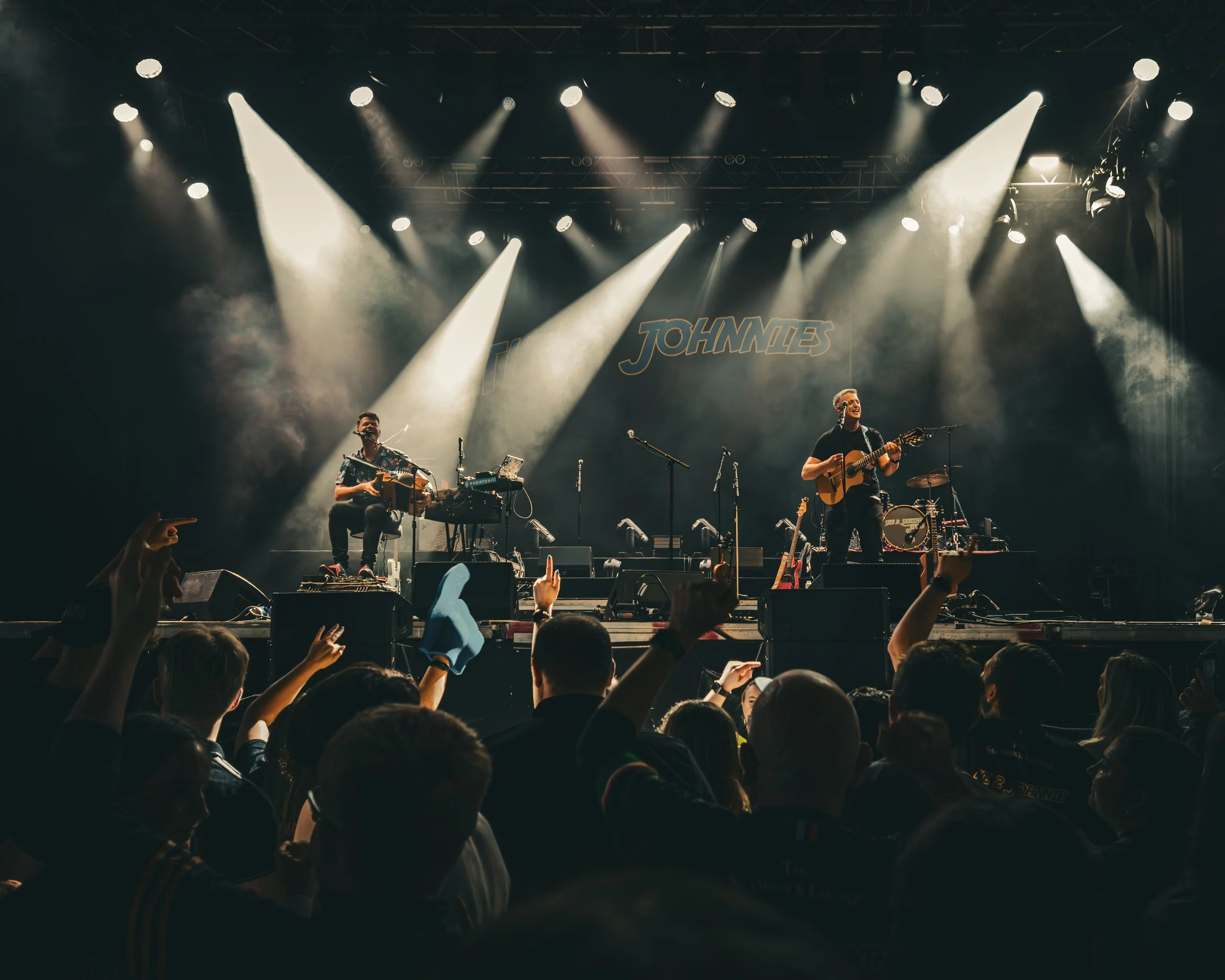 Musicians performing on stage at a concert, with audience members raising their hands and pointing towards the stage. The stage has the name "JOHNIES" displayed in the background.
