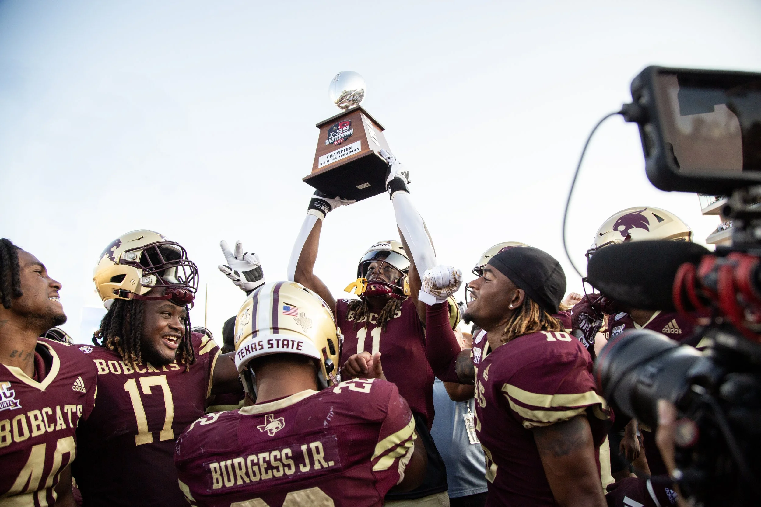 A Texas State University football player holds a trophy in the air celebrating a victory surrounded by his teammates.