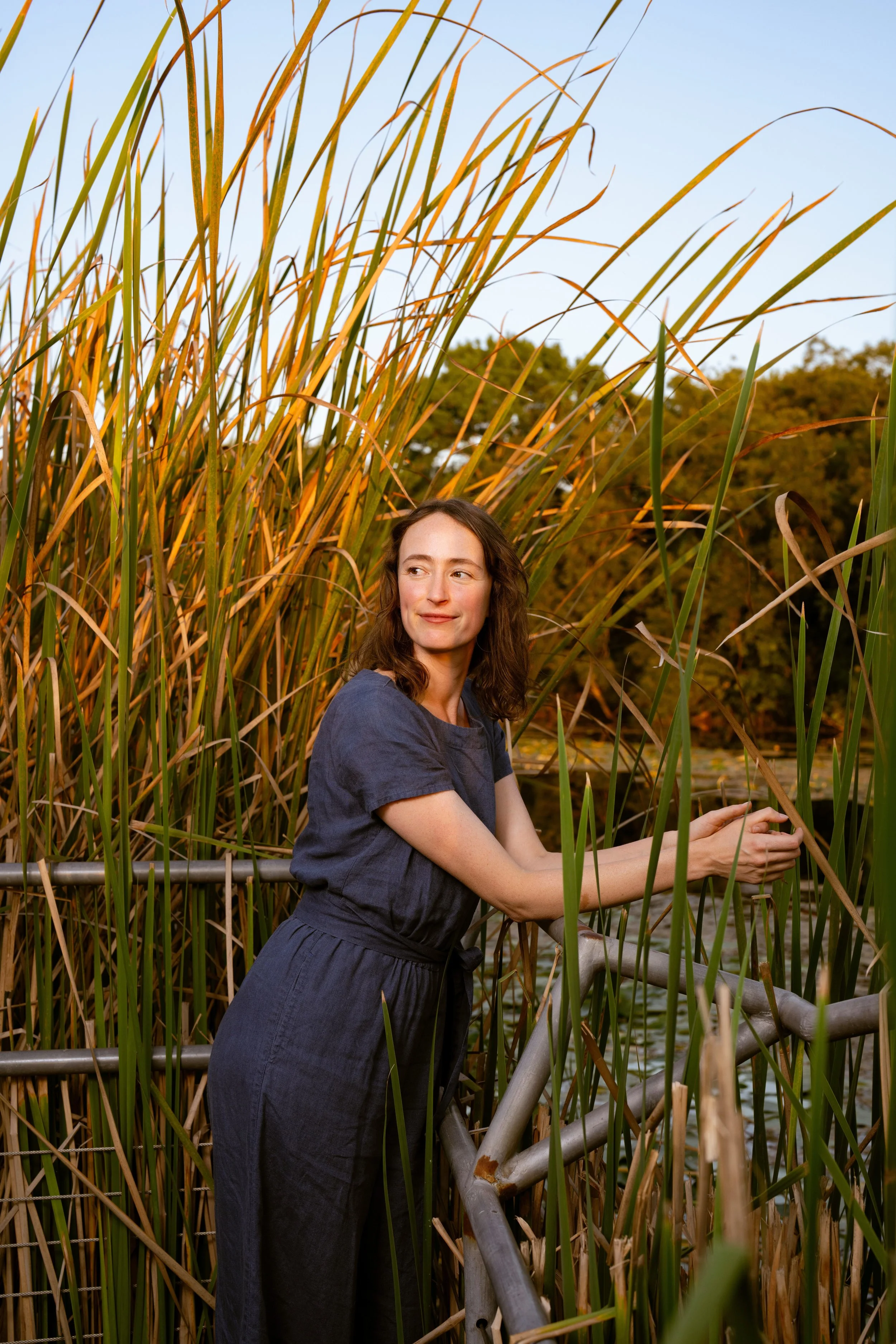 Bess Price leans over a railing surrounded by tall reed grass.