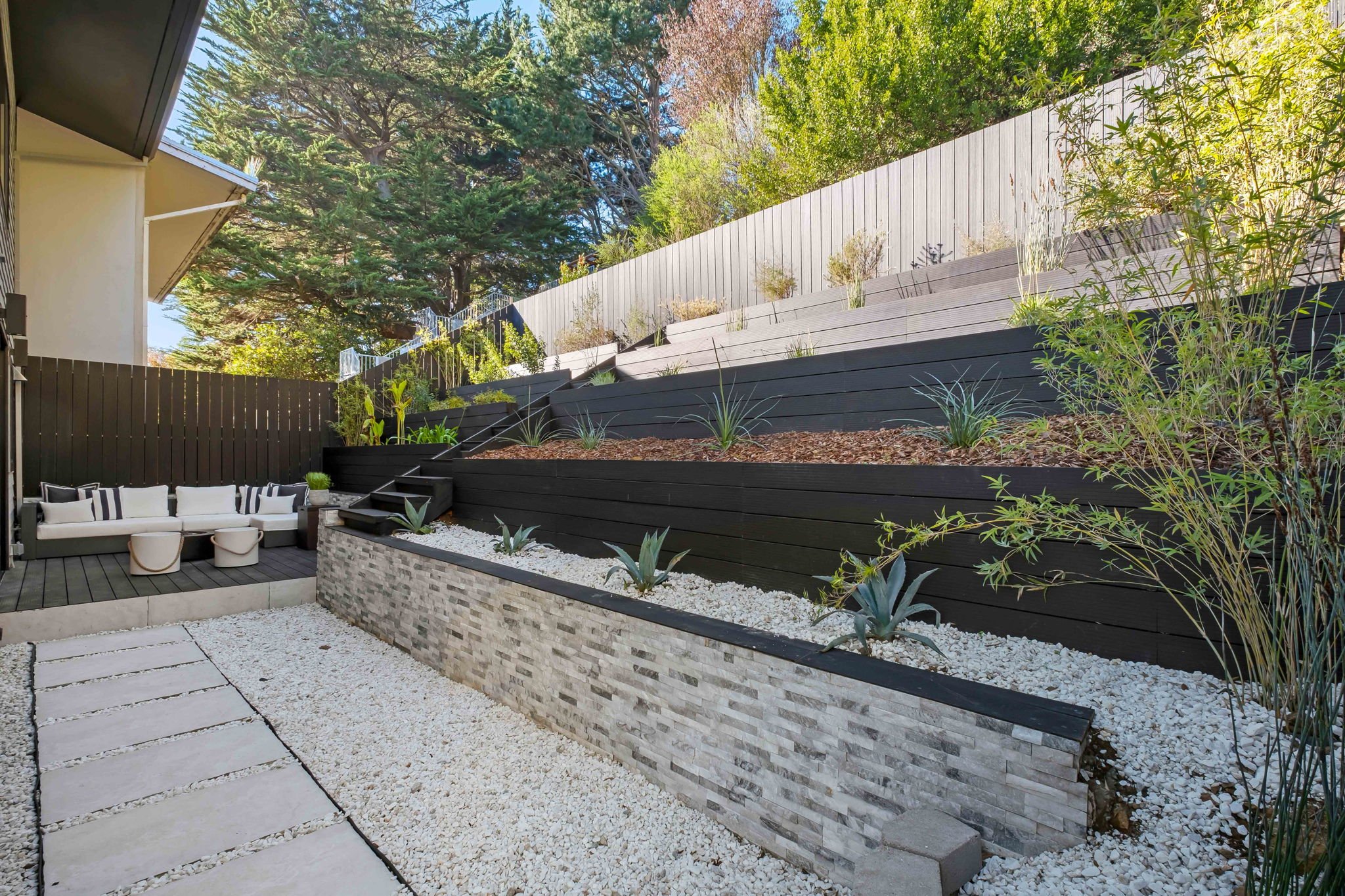 Modern backyard with tiered garden beds, black and white wood fencing, and outdoor seating area with white couches and small tables.