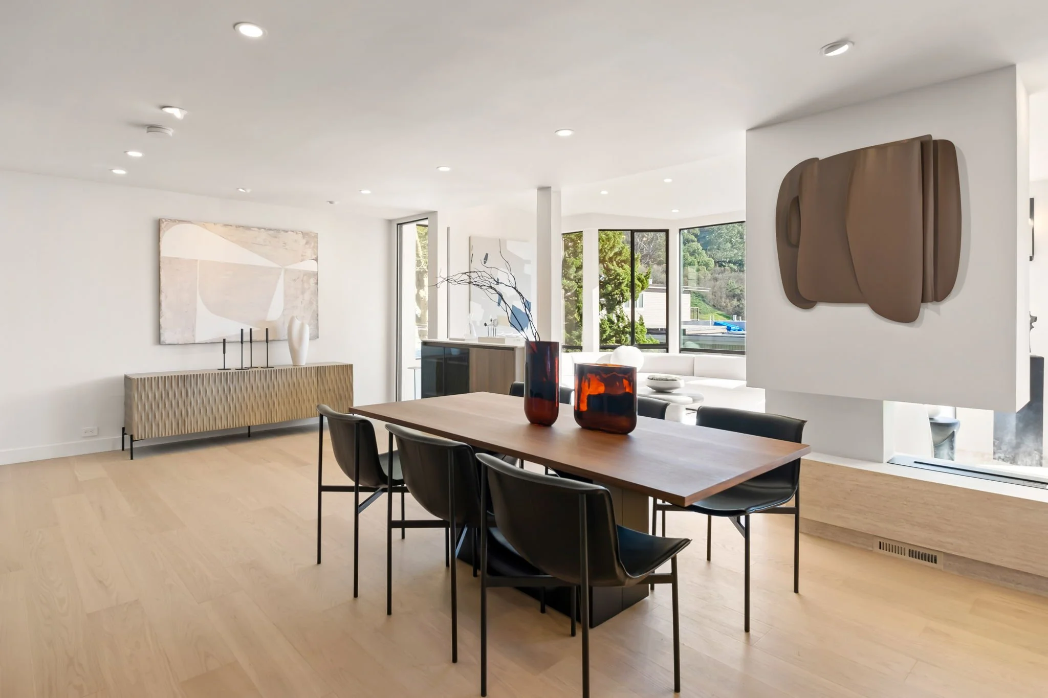 Modern dining room with wooden table, six black chairs, abstract wall art, contemporary sideboard, decorative vases, and large windows showing greenery outside.