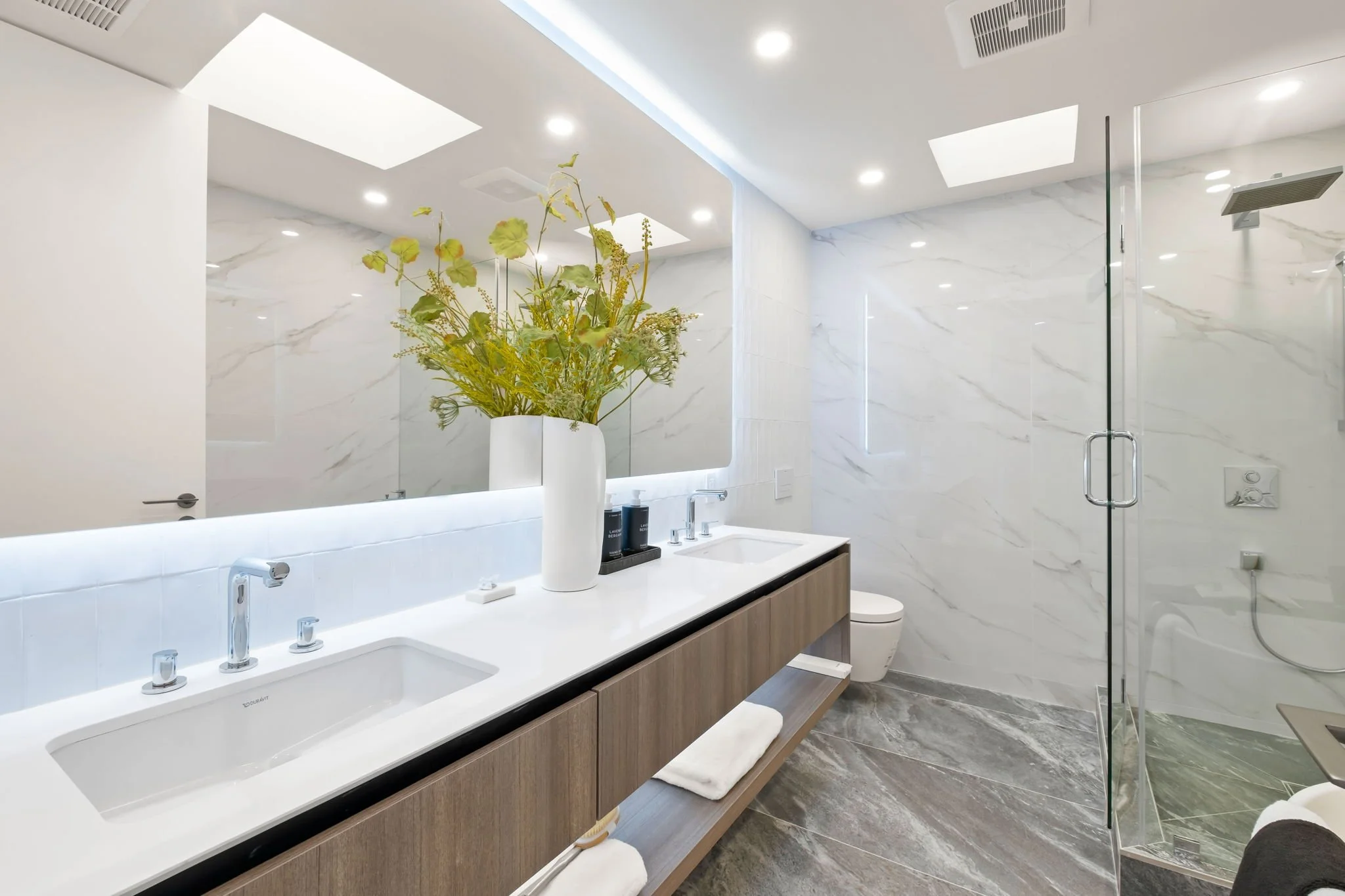 Modern bathroom with a double sink vanity, large mirror, decorative vase with flowers, marble walls, and a glass-enclosed shower.