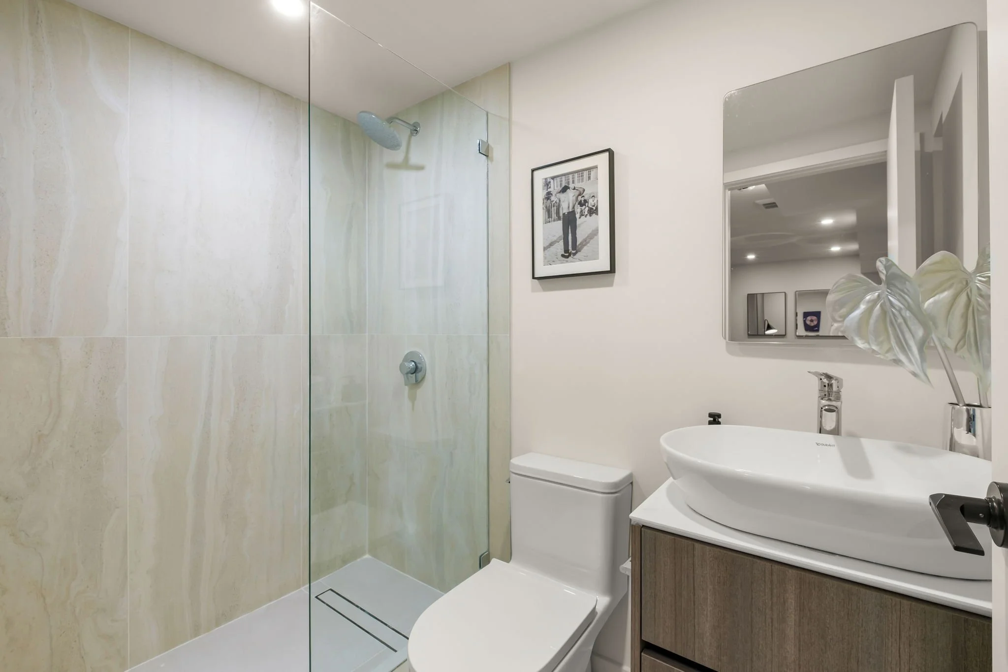 A modern bathroom with a glass-enclosed shower, a white toilet, a wooden vanity with a vessel sink, a large mirror, a black and white framed photo on the wall, and a door reflected in the mirror.
