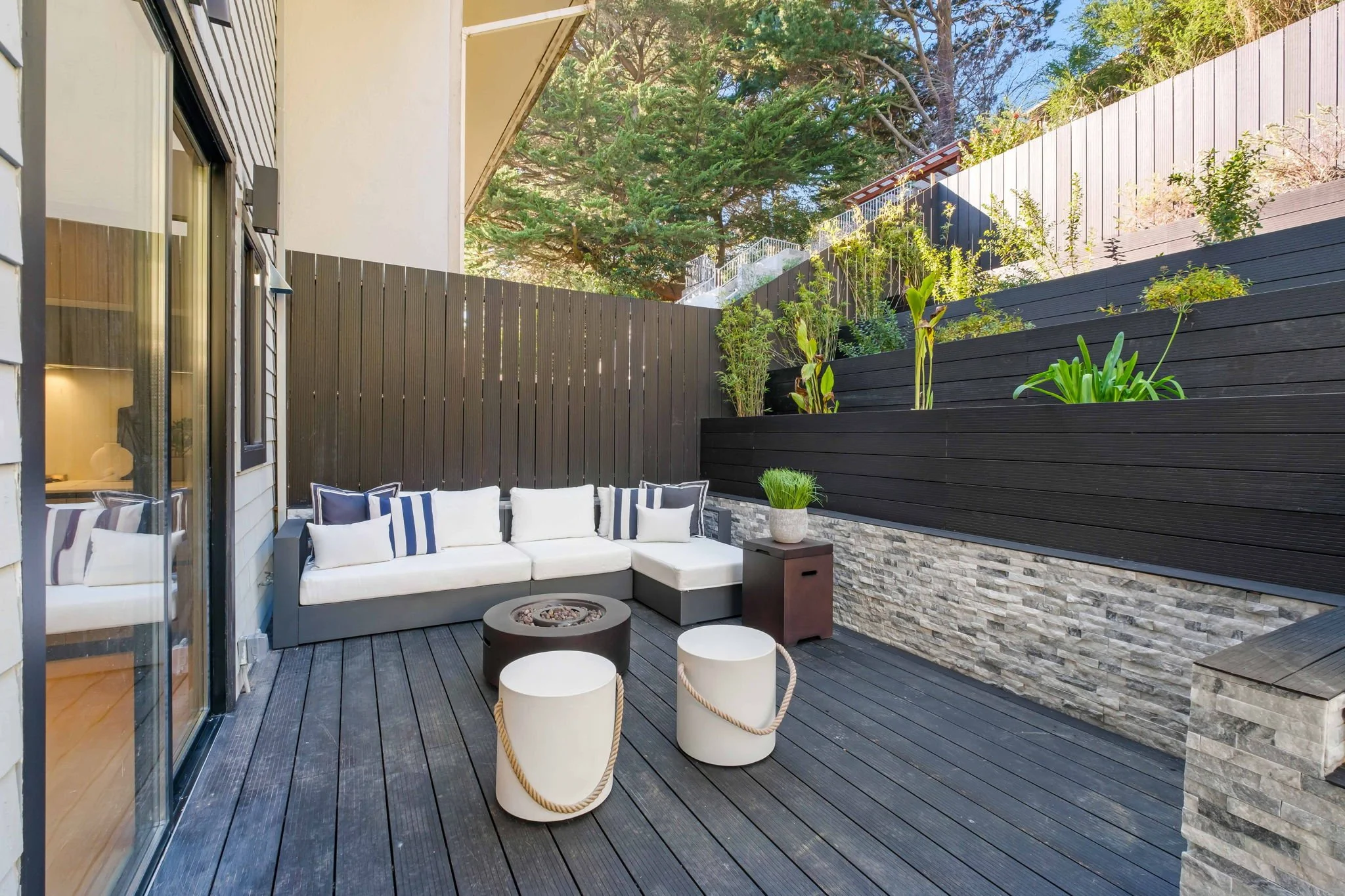 Modern outdoor patio with white sectional sofa, black firepit, two white stools with rope handles, potted plant, black wood fencing, and tiered garden with plants and trees.