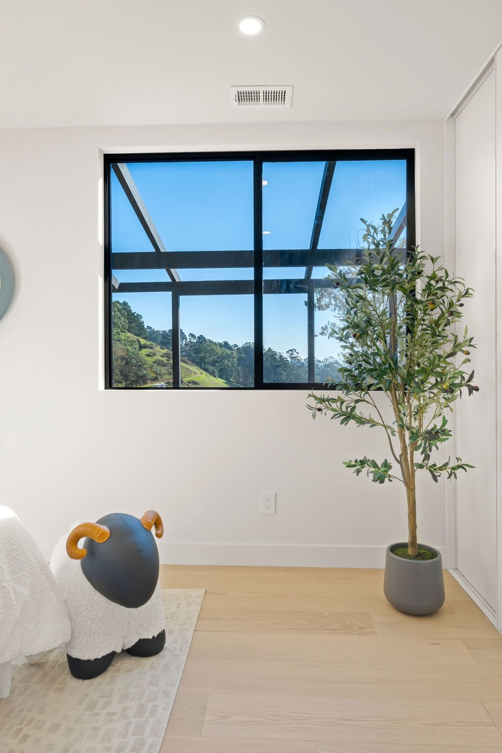 A minimalist room with a large black-framed window showing a view of a green landscape and blue sky. Inside, there is a tall potted plant with green leaves, a hidden bed with a sheep-shaped plush toy with ram horns, and a small beige rug on light woo