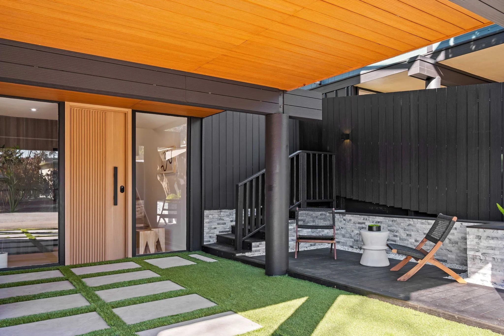Modern outdoor patio area with a small seating arrangement, wooden chairs, a white side table, and a grassy walkway leading to a glass sliding door of a contemporary house with black exterior walls and wooden accents.
