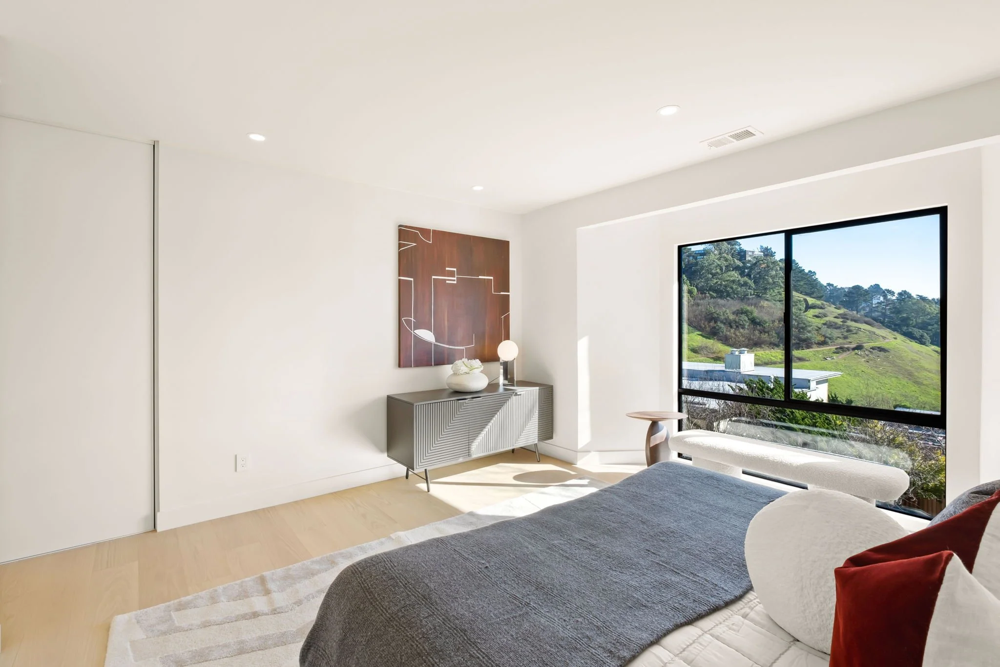 Bright, modern bedroom with large window overlooking green hills, bed with dark gray cover, white cylindrical pillow, and red pillows, grey sideboard with decorative objects, and abstract artwork on white wall.