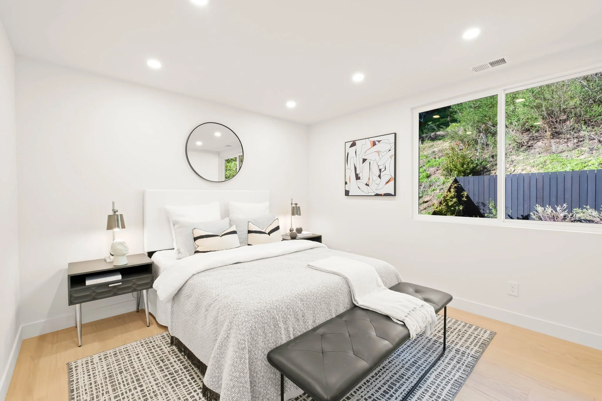 A modern bedroom with white walls, ceiling, and bedding, featuring black and neutral decor accents. The bed has four pillows, a gray blanket, and a white throw. Two bedside tables with lamps and decorative items. A round mirror on the wall and an abs