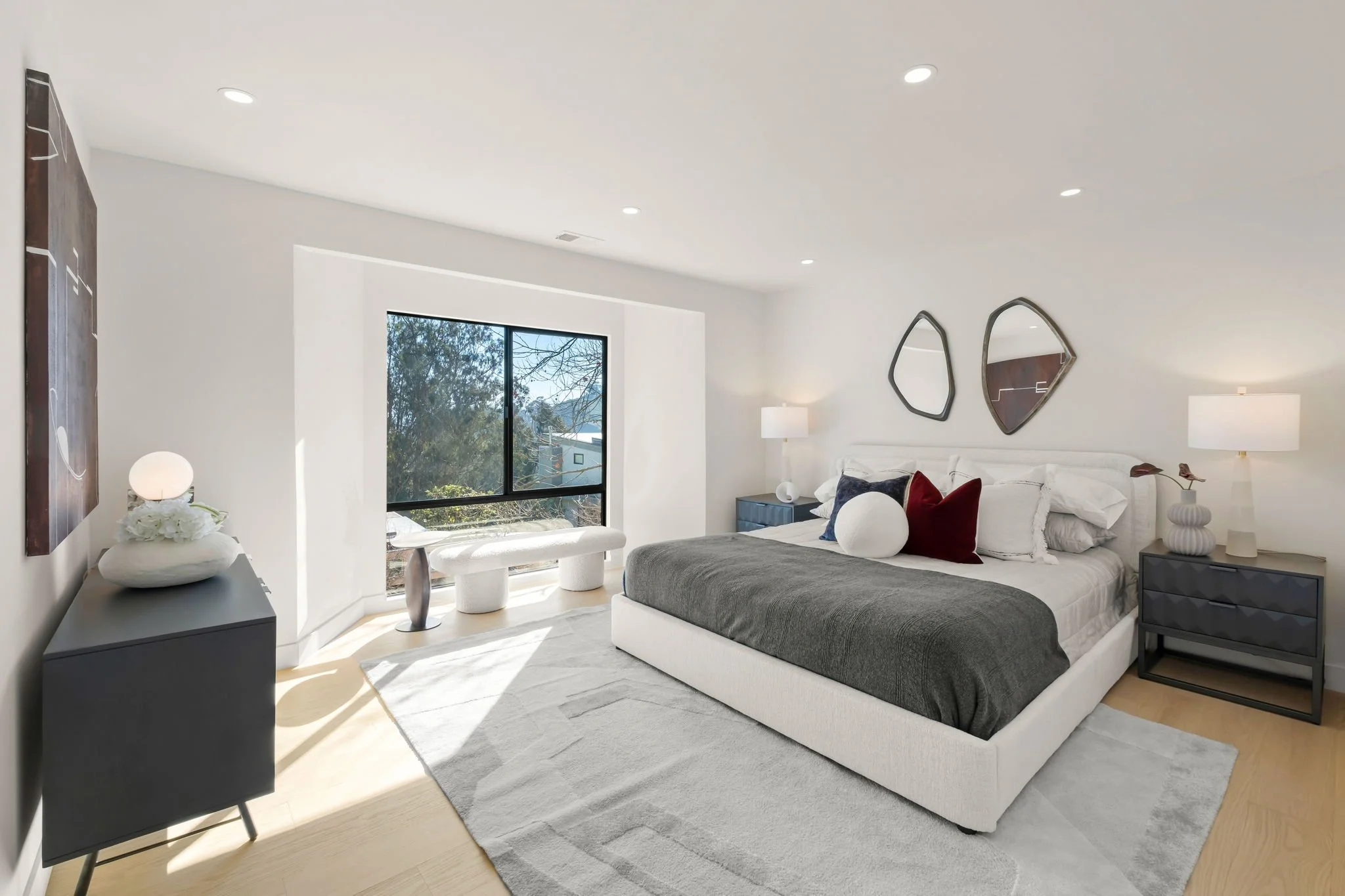 Modern bedroom with a large bed, white and navy pillows, gray blanket, and two black nightstands with lamps. Decorative mirrors on the wall and a window with a view of trees outside.